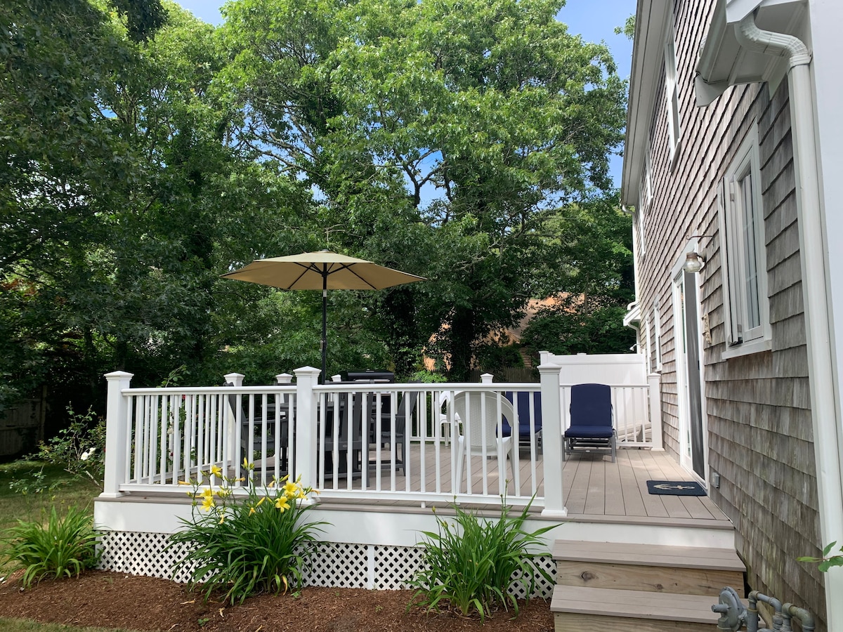 A spacious deck is visible, surrounded by lush greenery and tall trees. A large umbrella shades a seating area, which includes outdoor furniture. Bright flowers are planted along the deck's edge, enhancing the inviting outdoor space.
