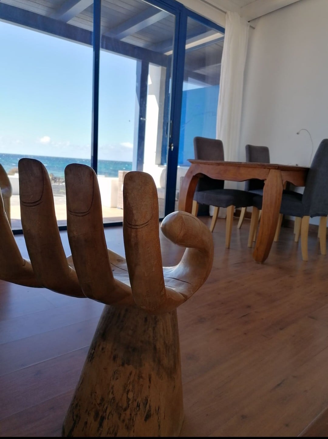 A unique wooden sculpture resembling a hand serves as decor in the foreground. Behind it, large glass doors offer a sweeping view of the Atlantic Ocean and light enters the space, highlighting a dining area with a wooden table and gray upholstered chairs.