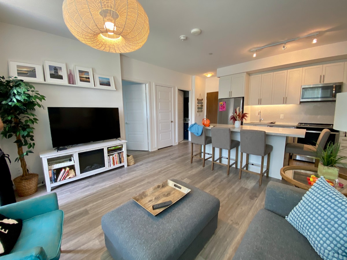 A modern living space features a large flat-screen TV on a white media console. An open-concept kitchen with stainless steel appliances and a spacious island is visible. Soft, natural lighting casts a welcoming glow on the furniture arrangement, including a comfortable seating area with decorative cushions.