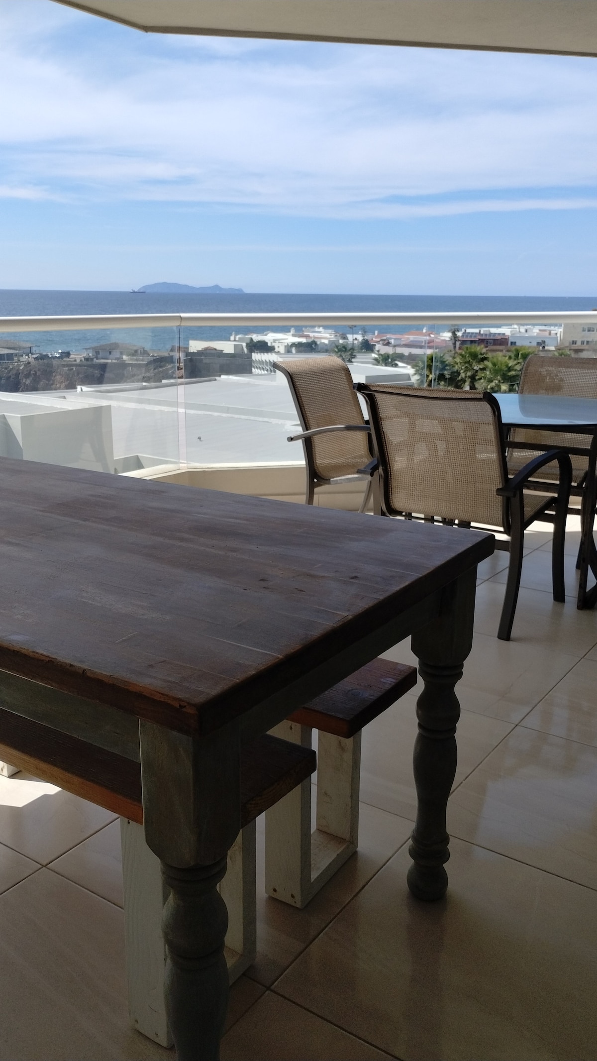 A wooden dining table is positioned on a shaded balcony, overlooking a distant view of the ocean and coastline. Metal chairs accompany the table, while a low glass railing provides an unobstructed sight of the water. The sky is filled with scattered clouds, enhancing the serene atmosphere.