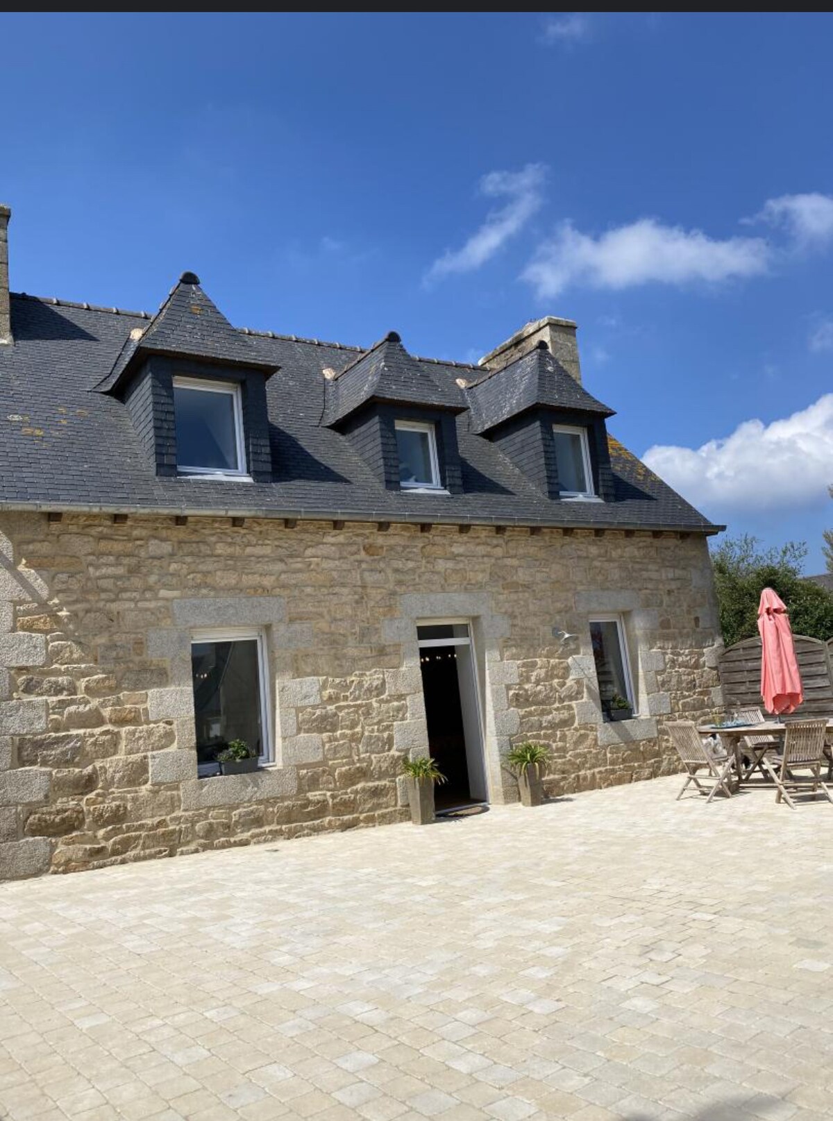 The exterior of a renovated stone house is captured under a clear blue sky. The façade features multiple gabled rooftops and two large windows. A small terrace with a table and chairs is visible, accompanied by a parasol providing shade.