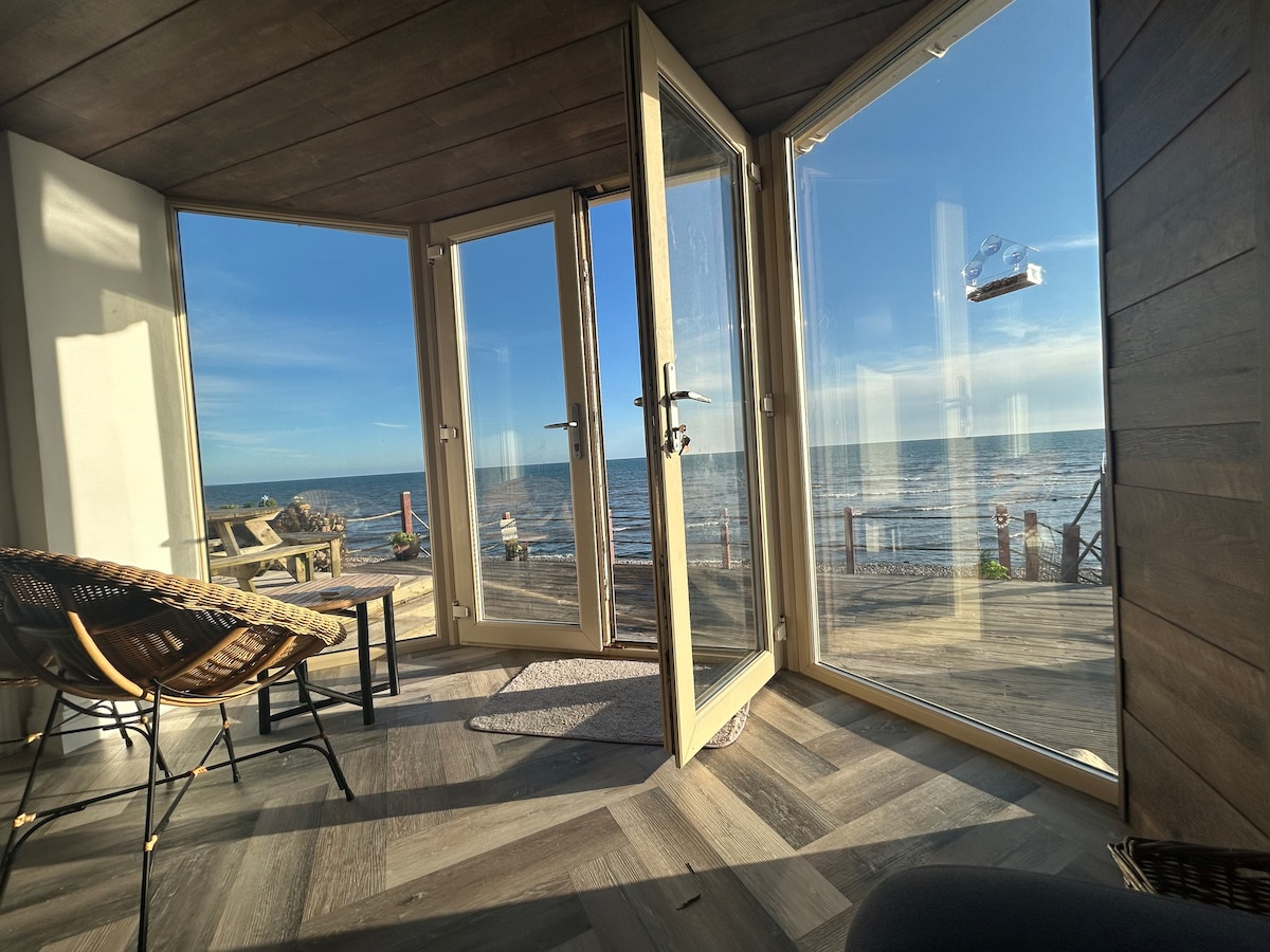An inviting corner features large glass doors that open to stunning sea views. Light wood flooring complements the natural hues of the decor. Two stylish chairs are positioned nearby, creating a comfortable area to enjoy the scenery and fresh sea air.
