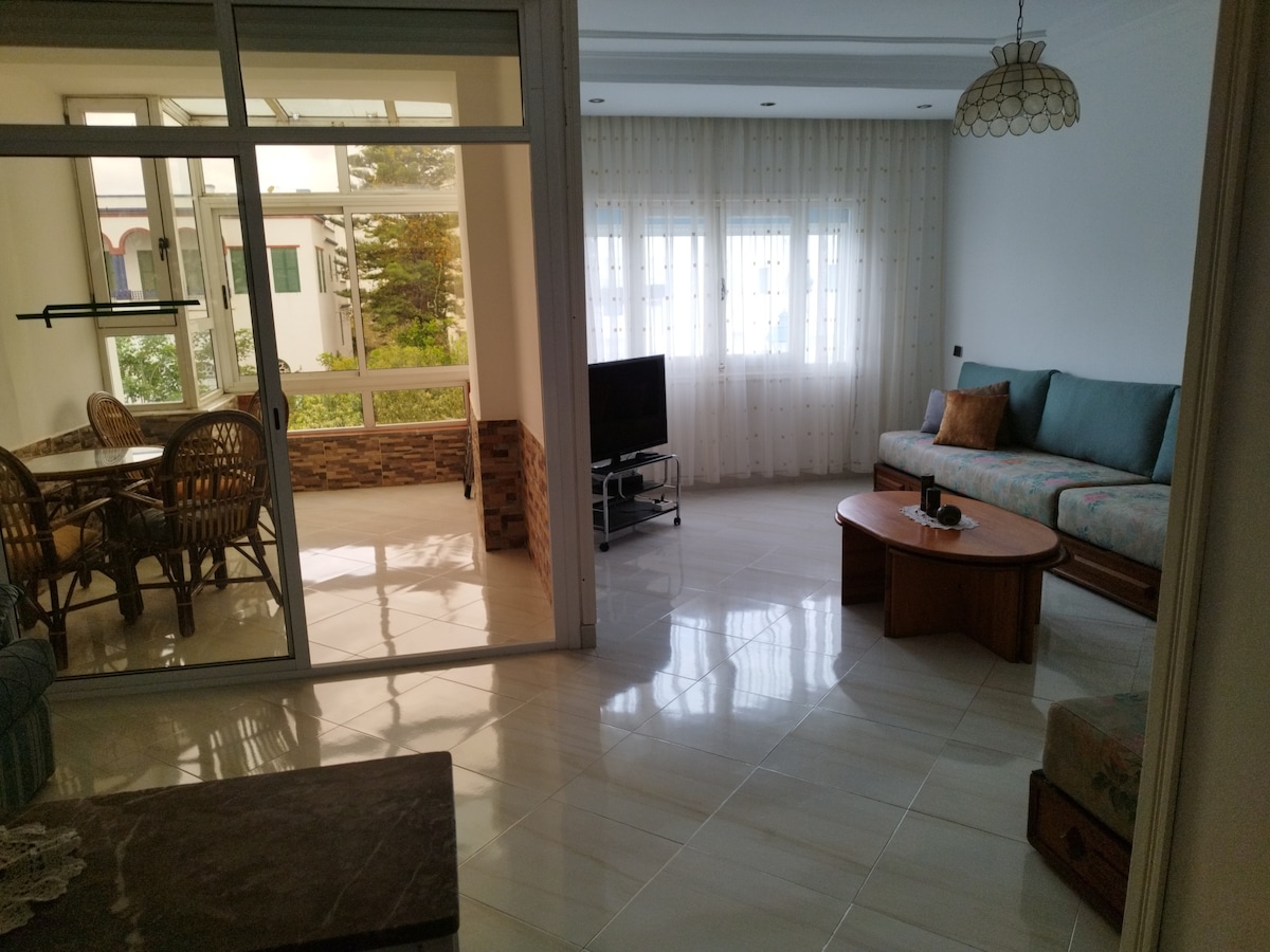 A well-lit living area features a spacious seating arrangement with a comfortable sofa and wooden coffee table. A television is positioned against a wall, while large windows allow natural light to fill the space. A cozy dining nook is visible through glass doors.