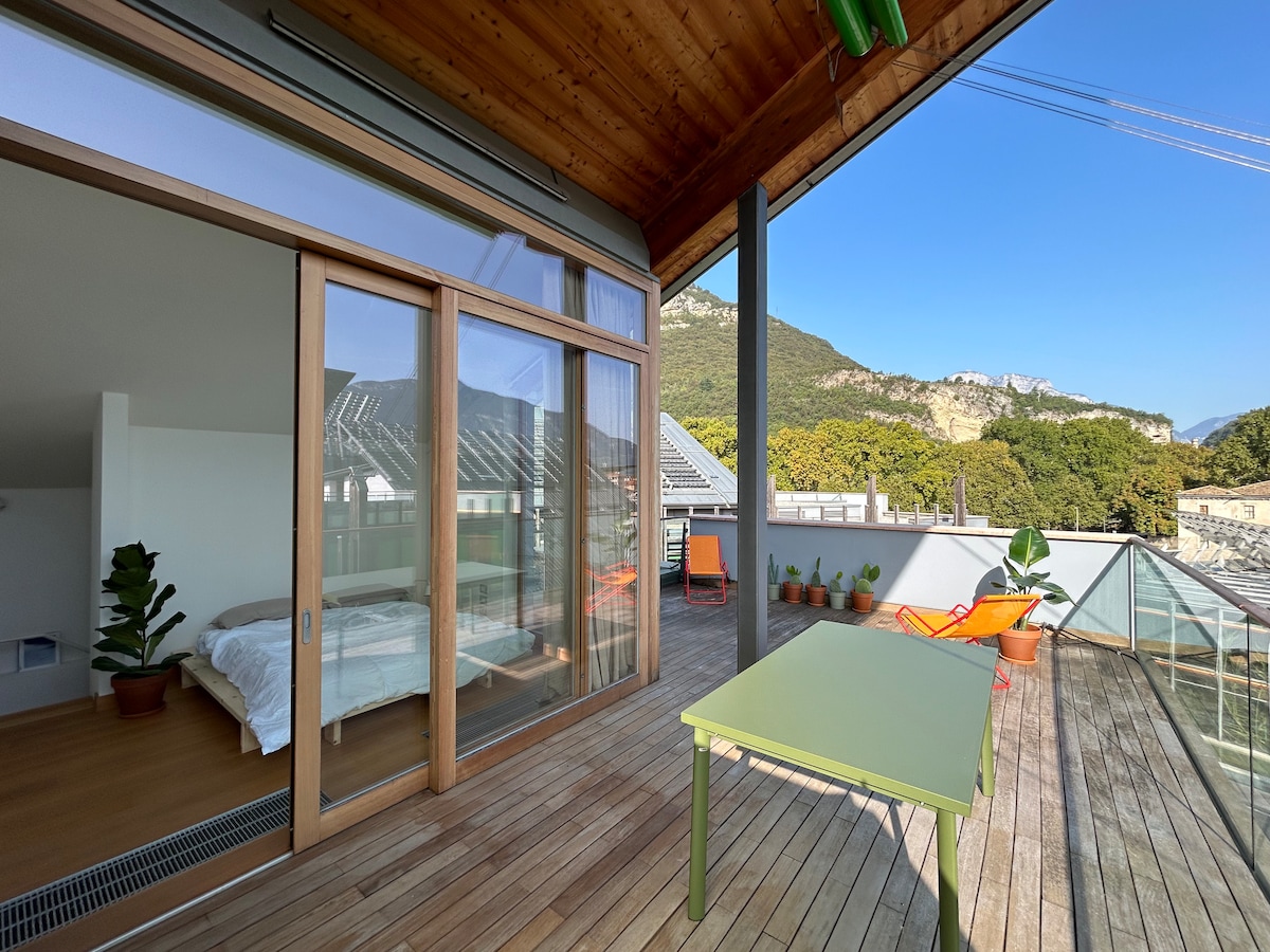 A spacious terrace features wooden decking, offering views of surrounding mountains and greenery. Large glass doors lead to an indoor area, revealing a bright room with a bed. Colorful outdoor seating options include an orange chair and a green table, creating a relaxed ambiance.