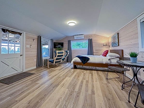 A spacious studio cottage features a comfortable king bed with crisp linens, situated near large windows. Cozy seating is provided by a pair of chairs and a small table, enhancing the welcoming interior. Light wood paneling and soft lighting create a relaxed atmosphere.