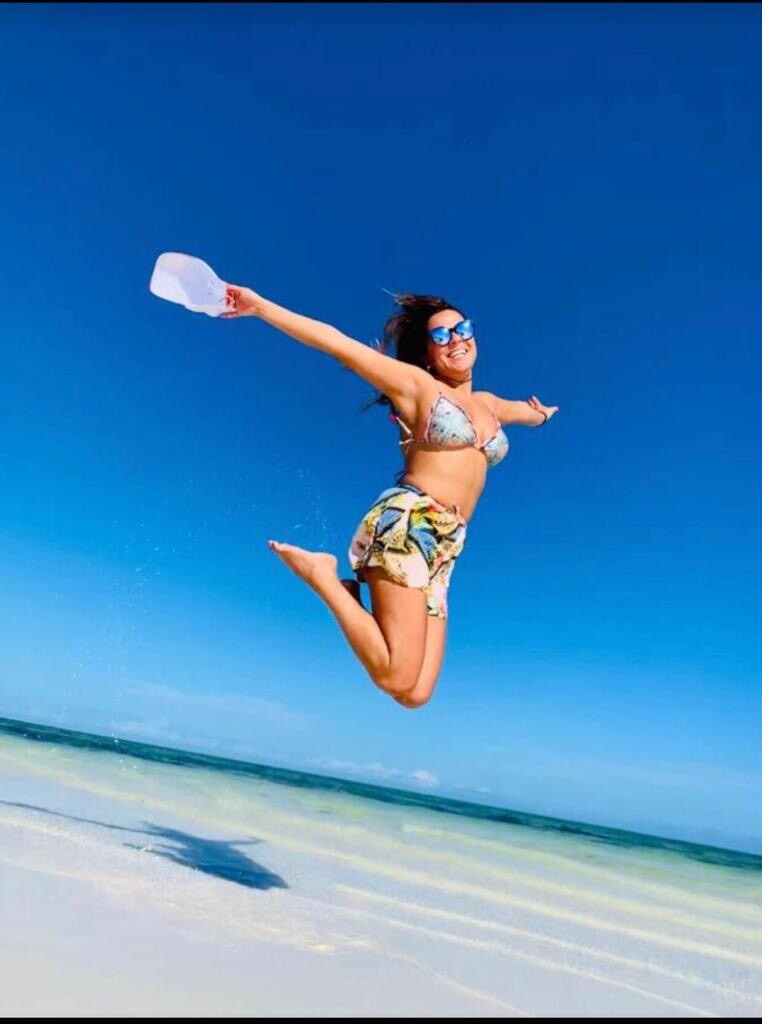 A person is seen leaping joyfully above a sandy beach, with the ocean waves gently lapping at the shore. The sky is clear and blue, creating a vibrant backdrop for this action shot.