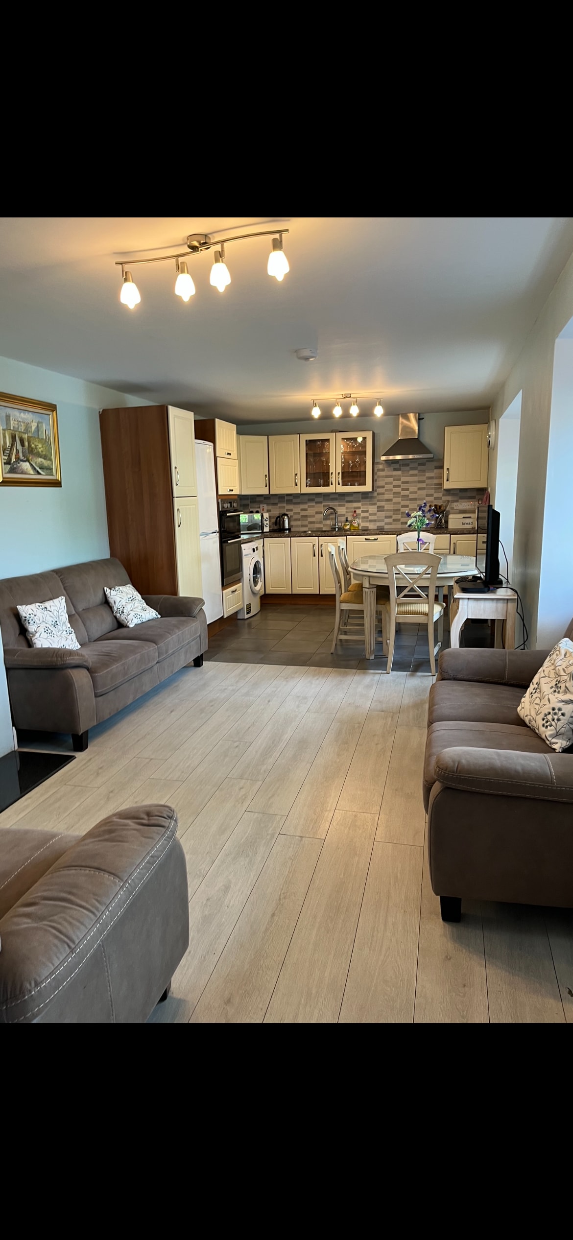 A spacious living area is presented, featuring two comfortable sofas with decorative pillows facing a dining table set for four. The kitchen area is visible in the background, equipped with cabinetry and modern appliances. Natural light brightens the room, enhancing the welcoming environment.