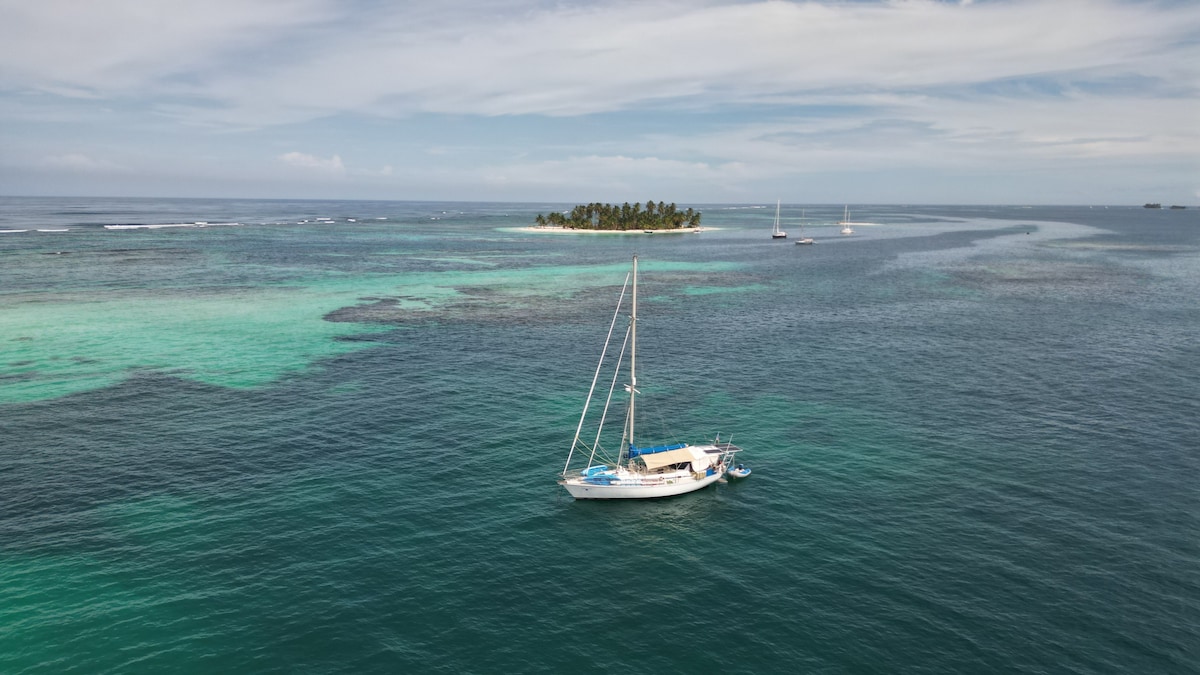 A sailboat is anchored in the clear turquoise waters, surrounded by lush greenery of a small island in the distance. The seascape displays varying shades of blue and green, with coral reefs visible beneath the surface, fostering a serene aquatic environment.