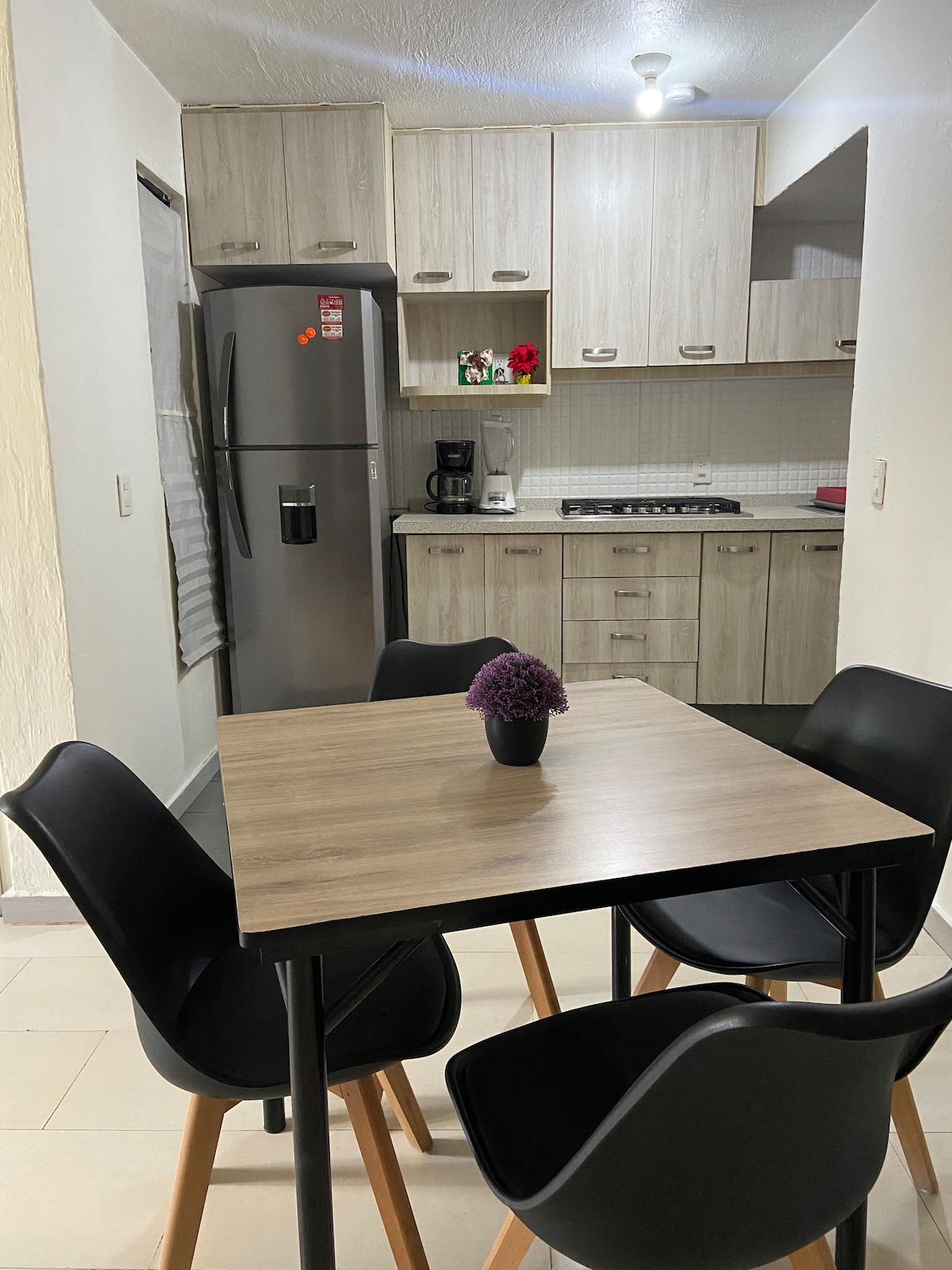 A modern kitchen space is depicted featuring light wood cabinetry and essential appliances. A dining table with four black chairs is positioned in the foreground. A small vase with purple flowers sits on the table, adding a touch of color.