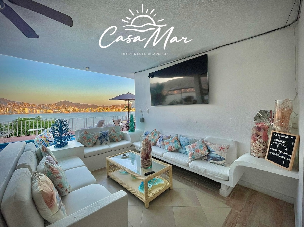A bright living area features white furniture accented with decorative cushions. A coffee table sits at the center, displaying seashells. The space is illuminated by natural light from large windows, offering views of the bay and surrounding hills.