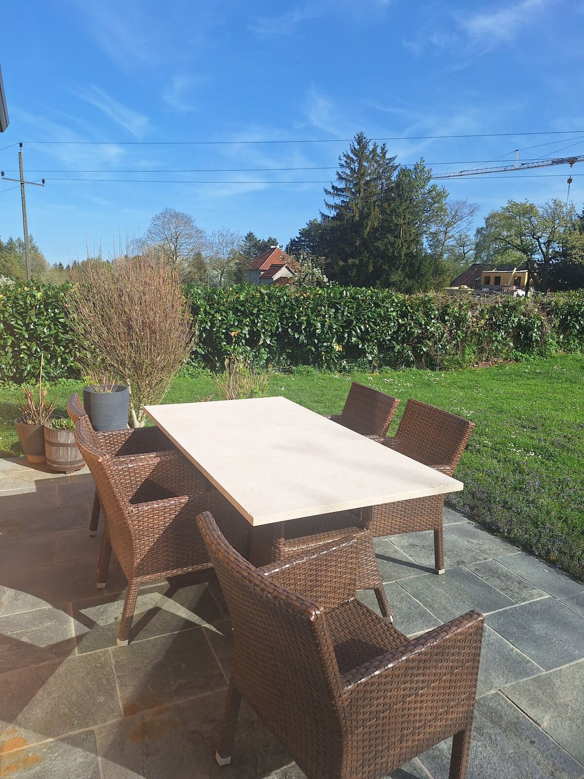 An outdoor seating area is presented with a stone table surrounded by four wicker chairs. Lush greenery is visible in the background, complementing a clear blue sky. Natural light enhances the inviting atmosphere of the space.