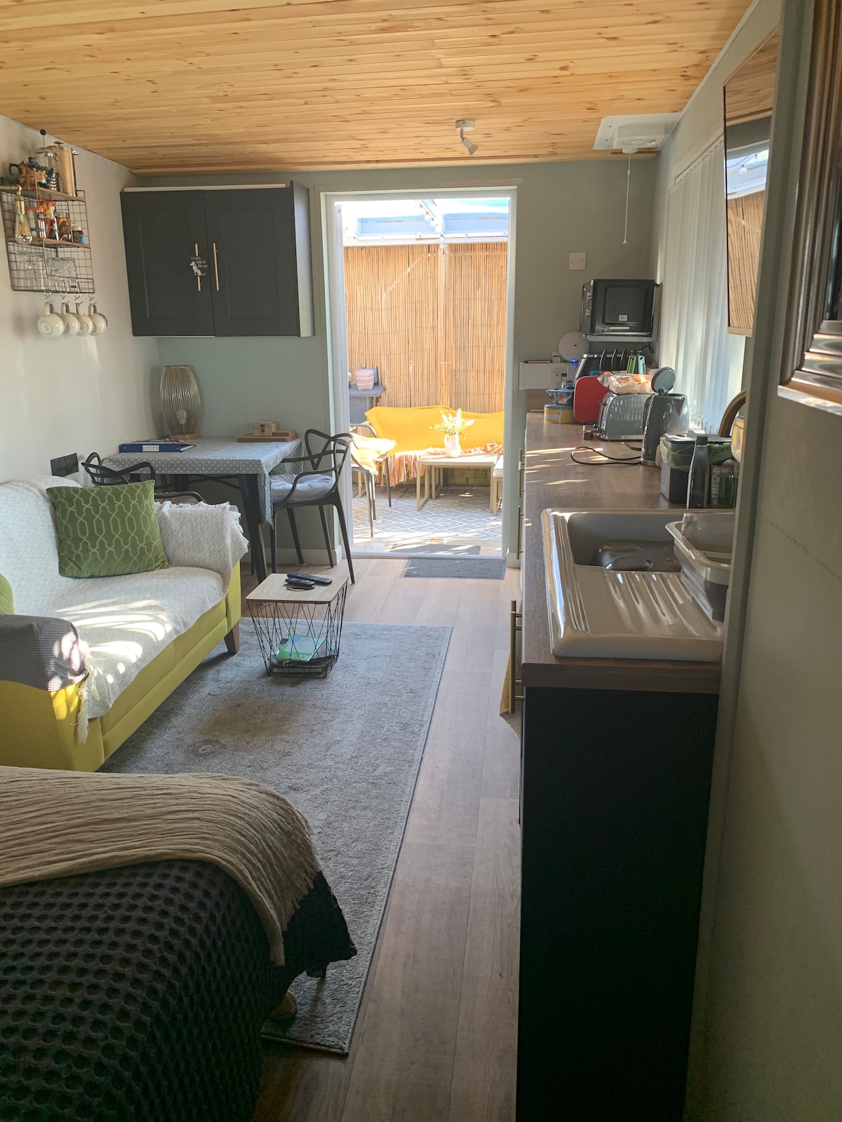 The interior of the Tarka Suite features a well-organized layout, showcasing a cozy seating area with a two-seater sofa and a dining table. A small kitchen area is visible, equipped with modern appliances. Natural light filters in through the door leading to a private outdoor space.