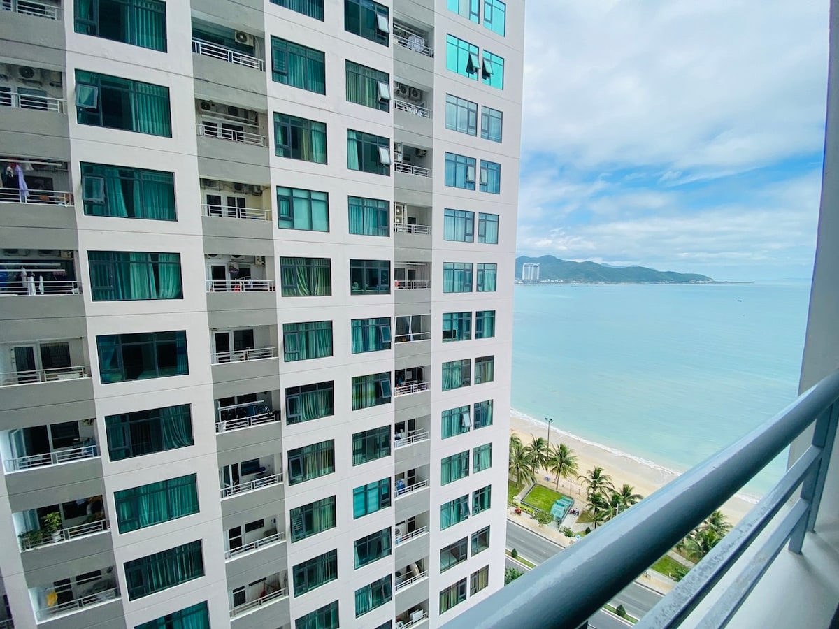 An exterior view of the OC2B Building is presented, showcasing its modern architecture. The sea can be seen in the background, with a glimpse of Hon Chong beach below. Floor-to-ceiling windows are visible, reflecting blue skies and an inviting environment.