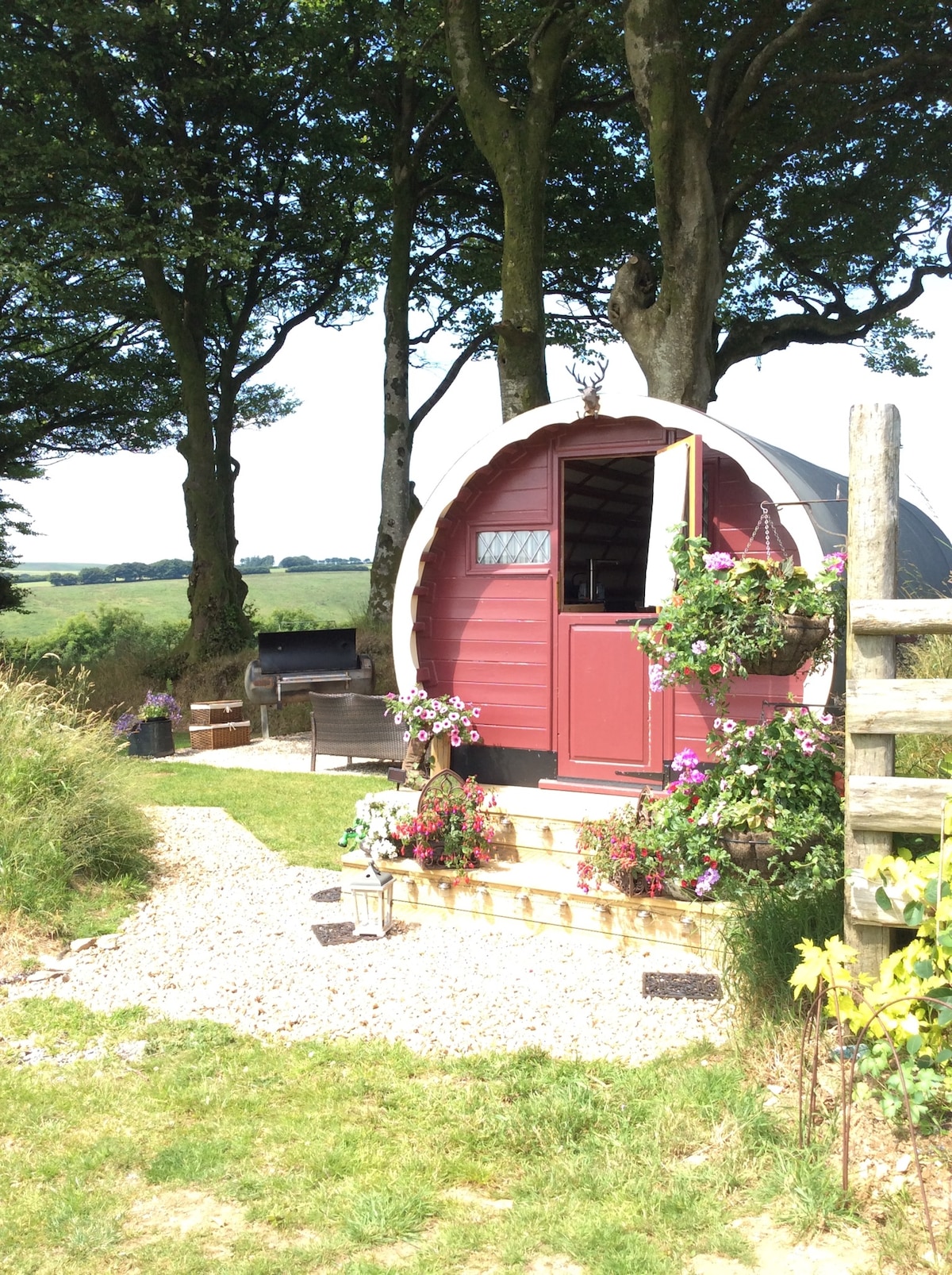 An inviting gypsy caravan is set among lush greenery, featuring a scalloped roof and vibrant flowers adorning its entrance. A small wooden deck leads up to the door, with a charcoal barbecue placed nearby, offering a serene view of the surrounding landscape.
