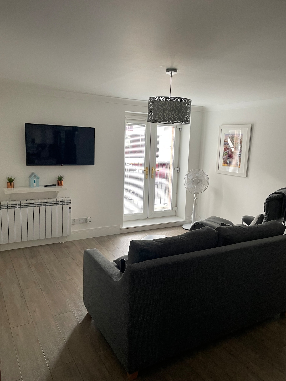 A light-filled living area features a comfortable gray sofa and a recliner chair. A television is mounted on the wall, providing entertainment options. Double doors open to an outdoor area, and a fan is positioned nearby for added comfort.