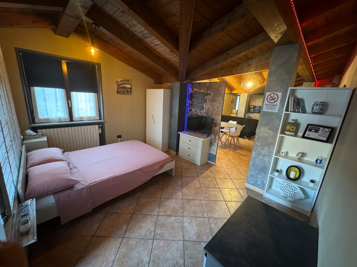 A bright sleeping area features a spacious bed with soft pink linens. Wooden beams are visible across the ceiling, while a window allows natural light to fill the room. Nearby, a dresser and a small shelving unit provide storage and décor.