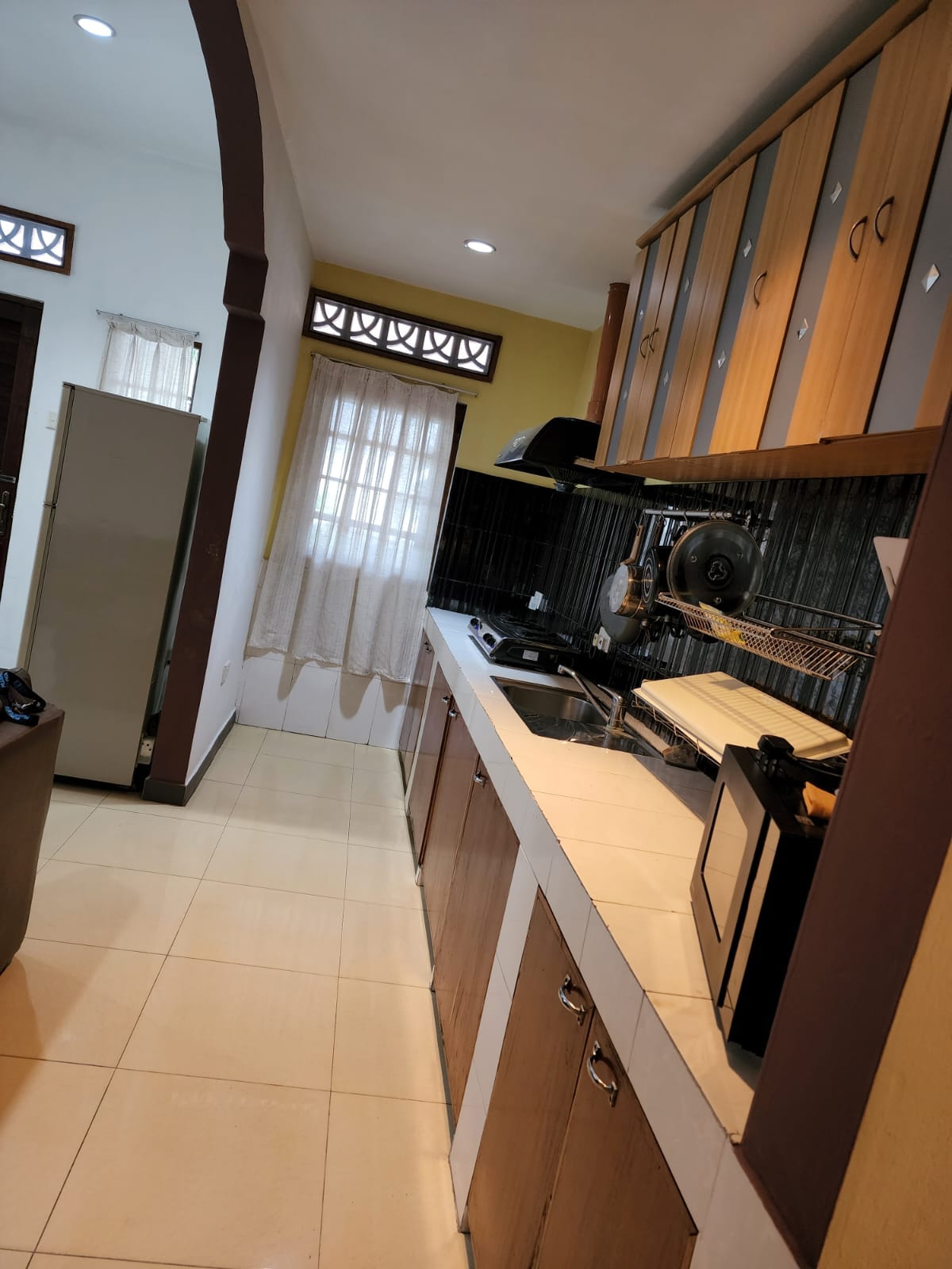 A full kitchenette is showcased, featuring wooden cabinetry and a clean countertop space. Essential appliances include a stove and a refrigerator. Natural light enters through a window covered by light curtains, illuminating the area. The tiled floor ensures easy maintenance.