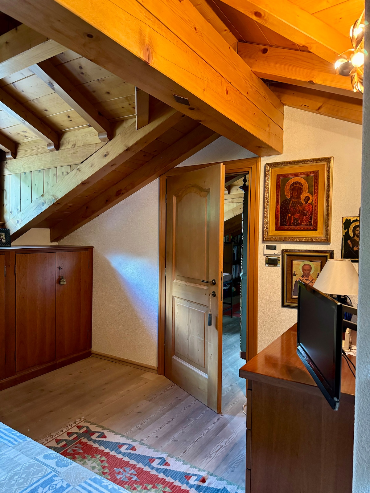 A warmly lit room features wooden beams across the ceiling, with a wooden door leading out. A dresser sits beside a small television, while decorative framed art pieces adorn the walls. Natural light filters in, enhancing the comfortable atmosphere.