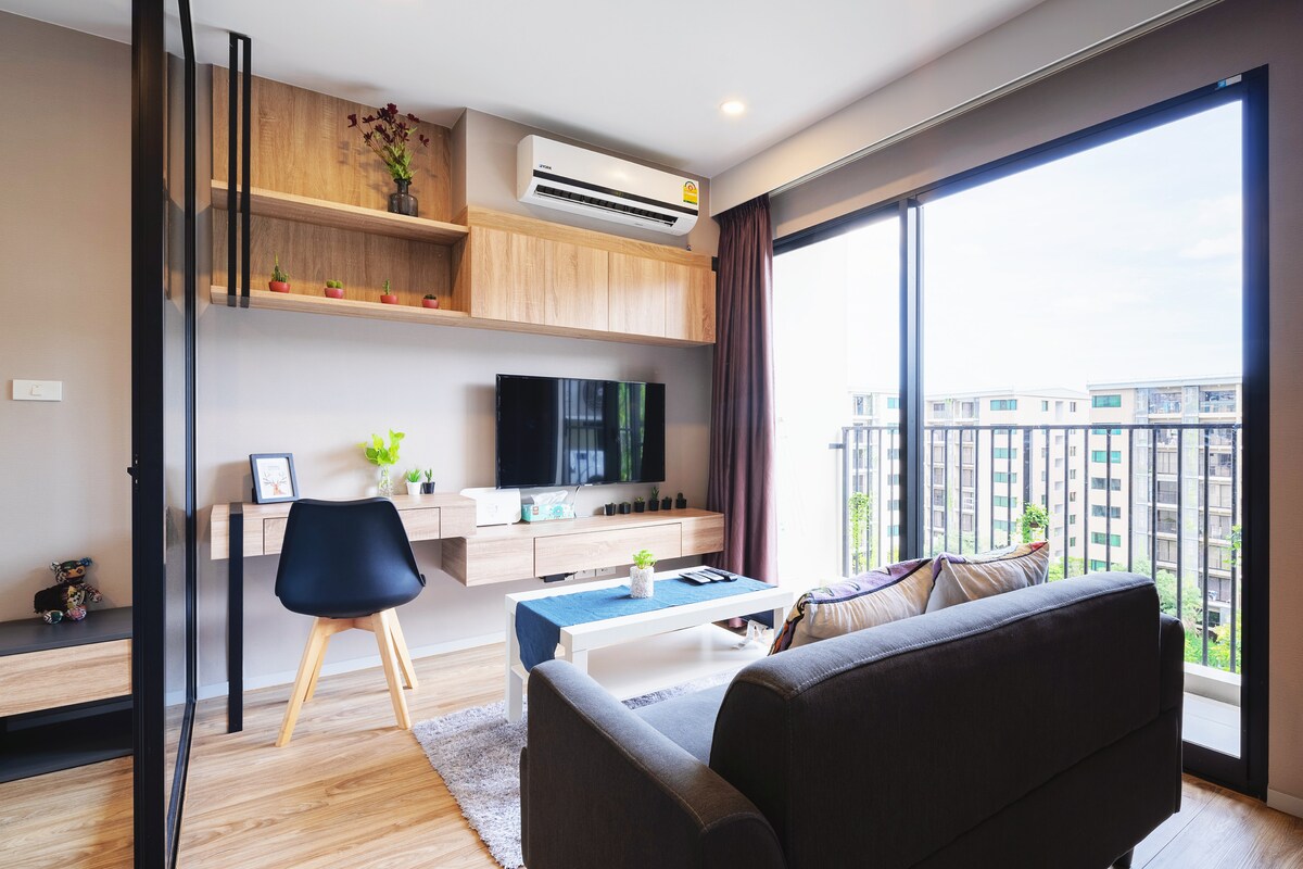 A modern living space features a comfortable sofa and a stylish chair. A wall-mounted television is positioned above a sleek wooden console. Large windows provide ample natural light and an outdoor view, while decorative shelf plants add a touch of greenery.