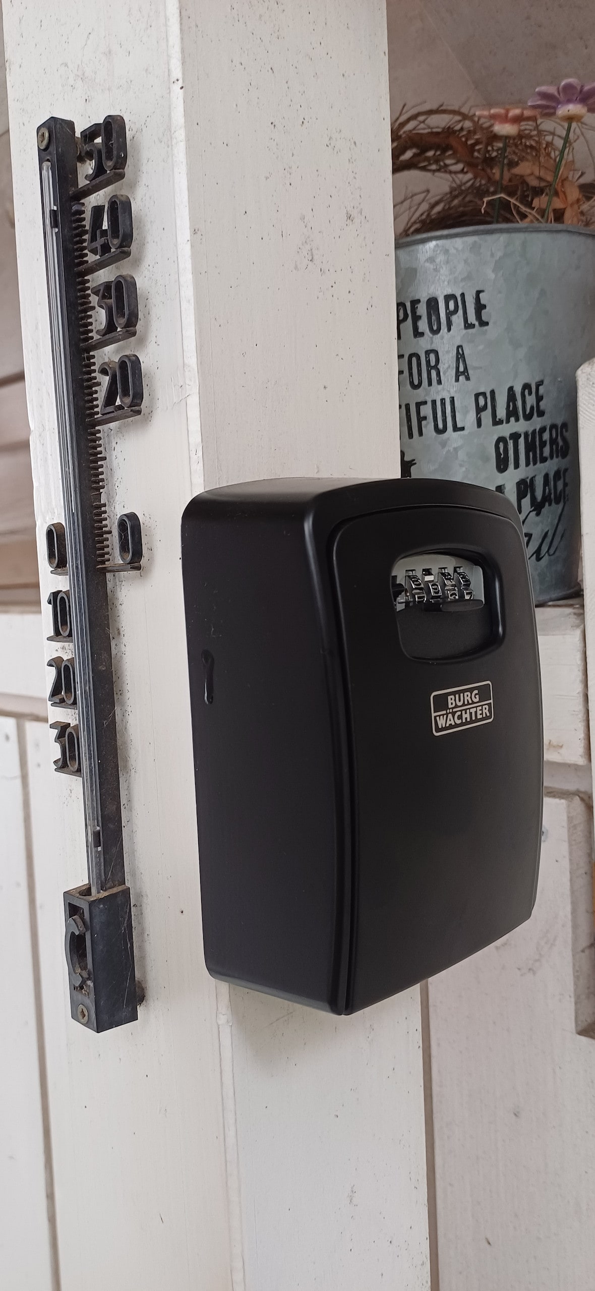 A black electronic lockbox is mounted on a white wooden wall. A measuring tape is attached alongside the lockbox, providing a clear view of the tool. The simplicity of the features emphasizes practicality and security.
