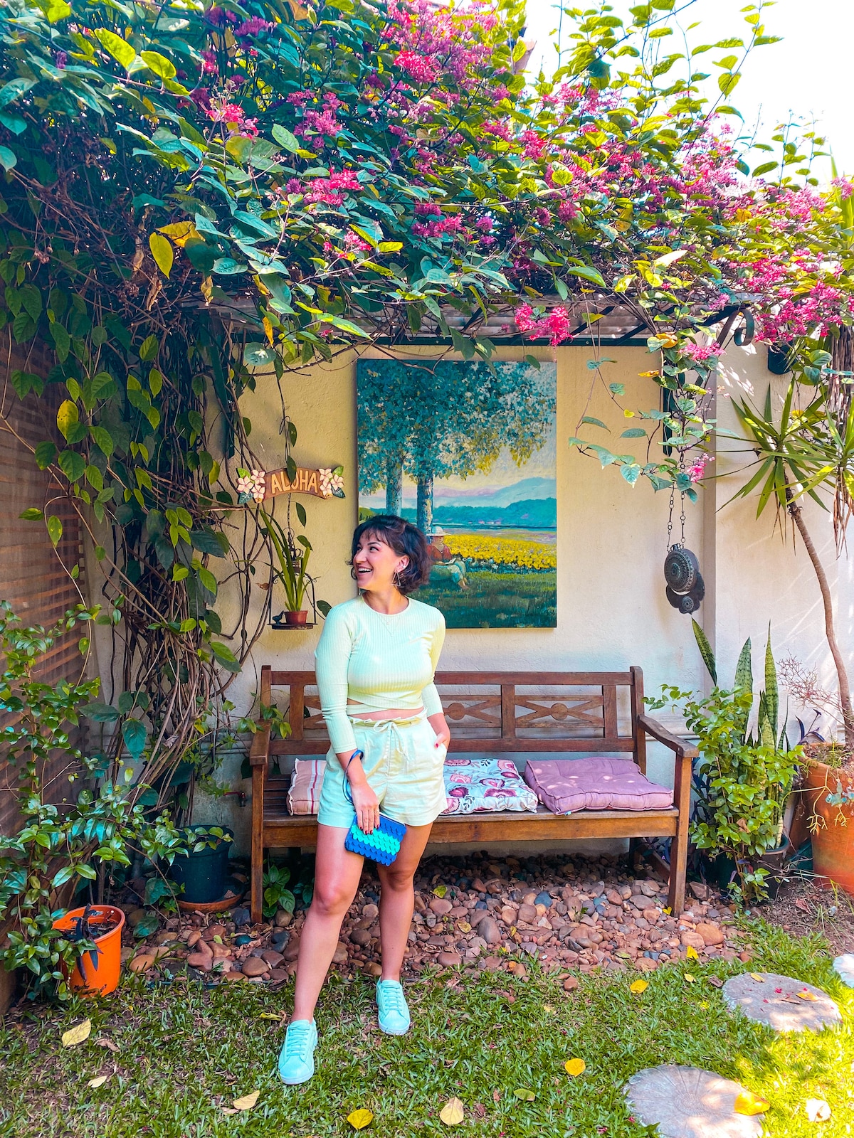 A serene garden area is highlighted, featuring a wooden bench adorned with cushions. Lush greenery and vibrant flowers frame the space, while a colorful painting of a landscape is mounted on the wall, adding a touch of artistry to the natural setting.