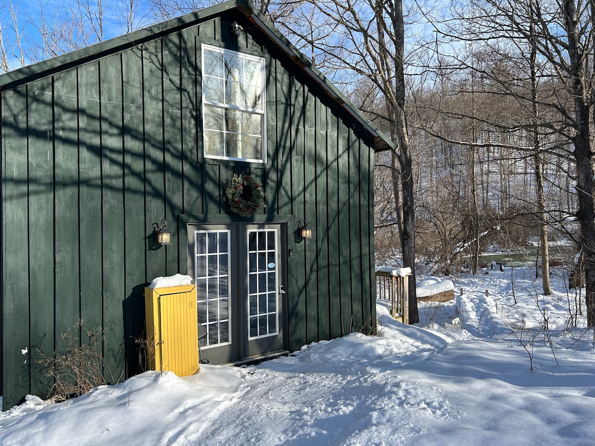 The exterior of the cottage features rustic green wooden siding with large windows. A bright yellow utility bin adds a pop of color beside the double doors. Snow covers the ground, and trees with bare branches are visible in the background, enhancing the serene winter setting.