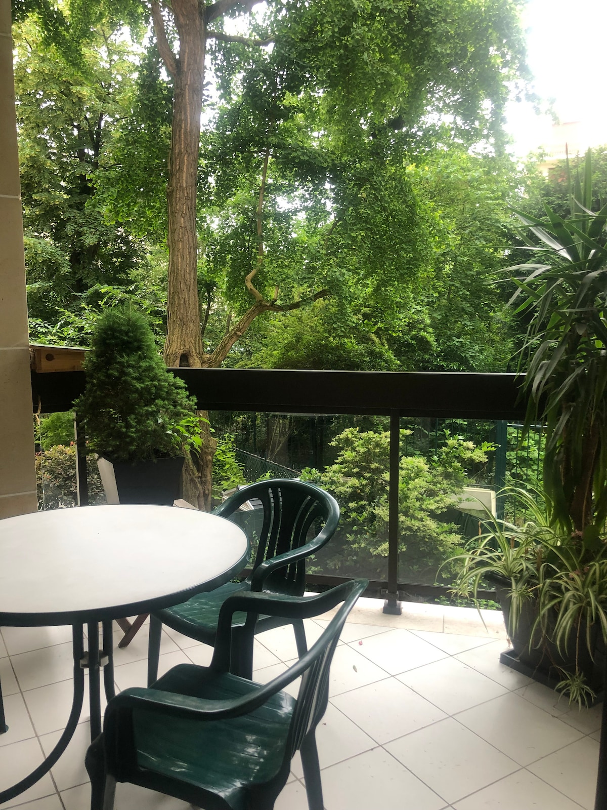 A private balcony is presented with a round table and three green chairs. Lush greenery surrounds the area, providing a tranquil view of vibrant trees and plants. The space is enhanced by potted plants, creating a serene outdoor setting.