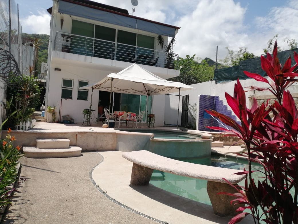 A modern single-level home is featured with a spacious outdoor area, including a well-maintained garden, a swimming pool, and a jacuzzi. A shaded seating area is positioned nearby, surrounded by greenery and vibrant plants, providing a serene environment for relaxation.