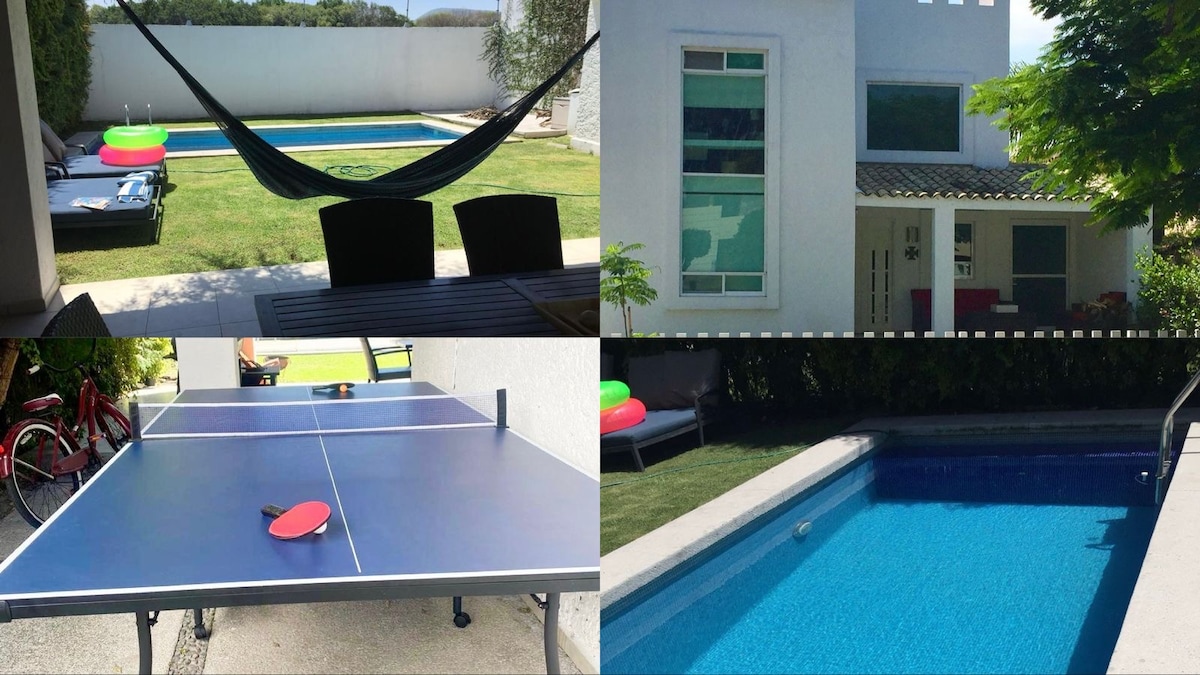 The outdoor area features a green lawn with a swimming pool bordered by a patio. A table tennis table is set up nearby, while a hammock is positioned for relaxation. Bright inflatable floaties add a splash of color to the pool, inviting leisure activities.