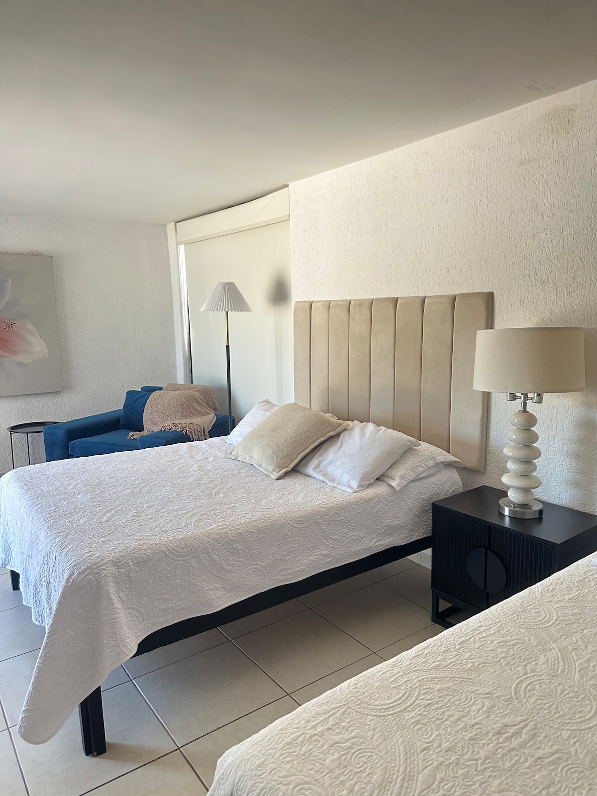 A light and airy bedroom is presented, featuring two spacious beds with white bedcovers. A blue armchair adds a touch of color, complemented by a standing lamp and a bedside table with a decorative lamp. Natural light enters through large windows.