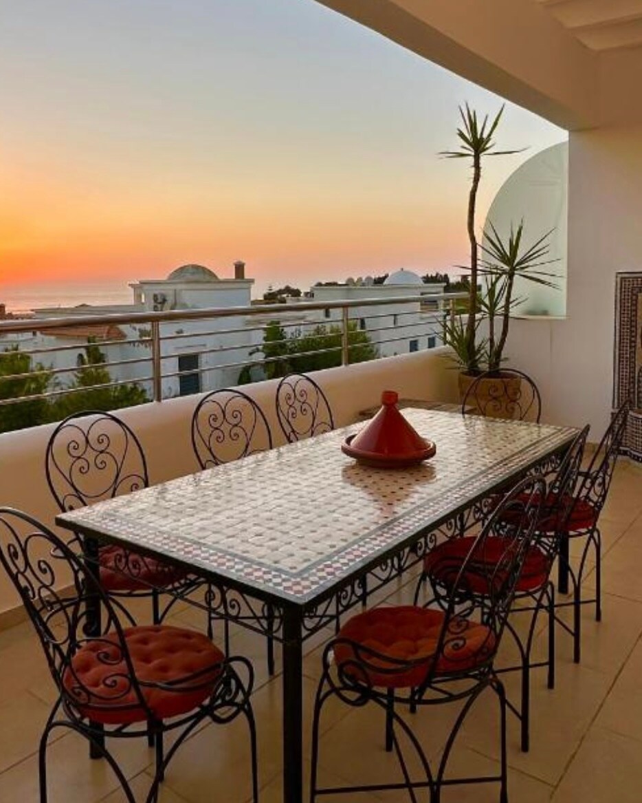An outdoor terrace features a large table surrounded by eight ornate chairs. A traditional Moroccan tagine is placed at the center of the table. The backdrop showcases a vibrant sunset with hues of orange and pink, along with views of the surrounding architecture and greenery.
