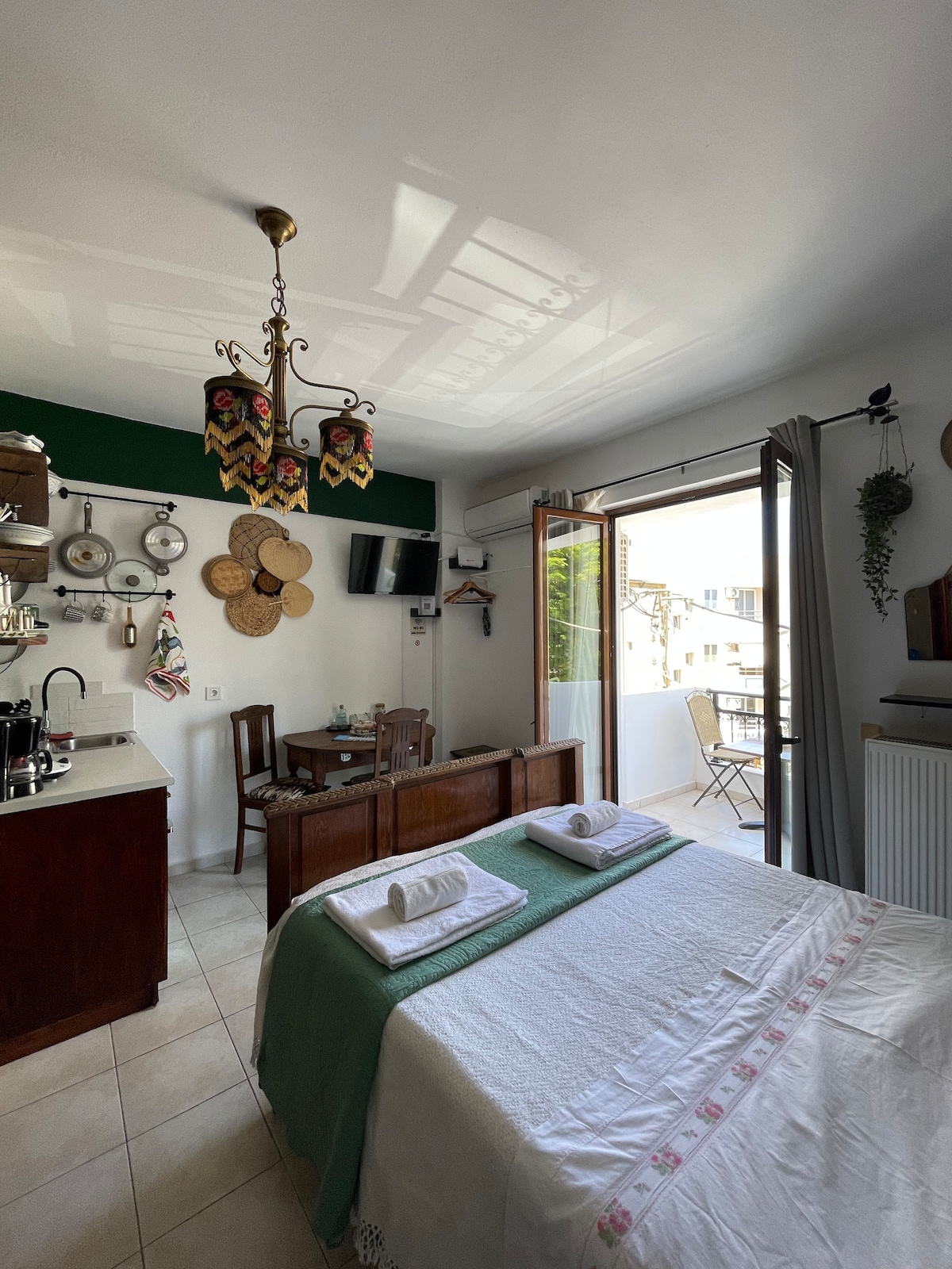 A well-equipped studio features a spacious bed with neatly arranged towels on top. A table for dining is positioned near the window that offers views of the surroundings. Decorative items add character, while a small balcony invites natural light into the living area.