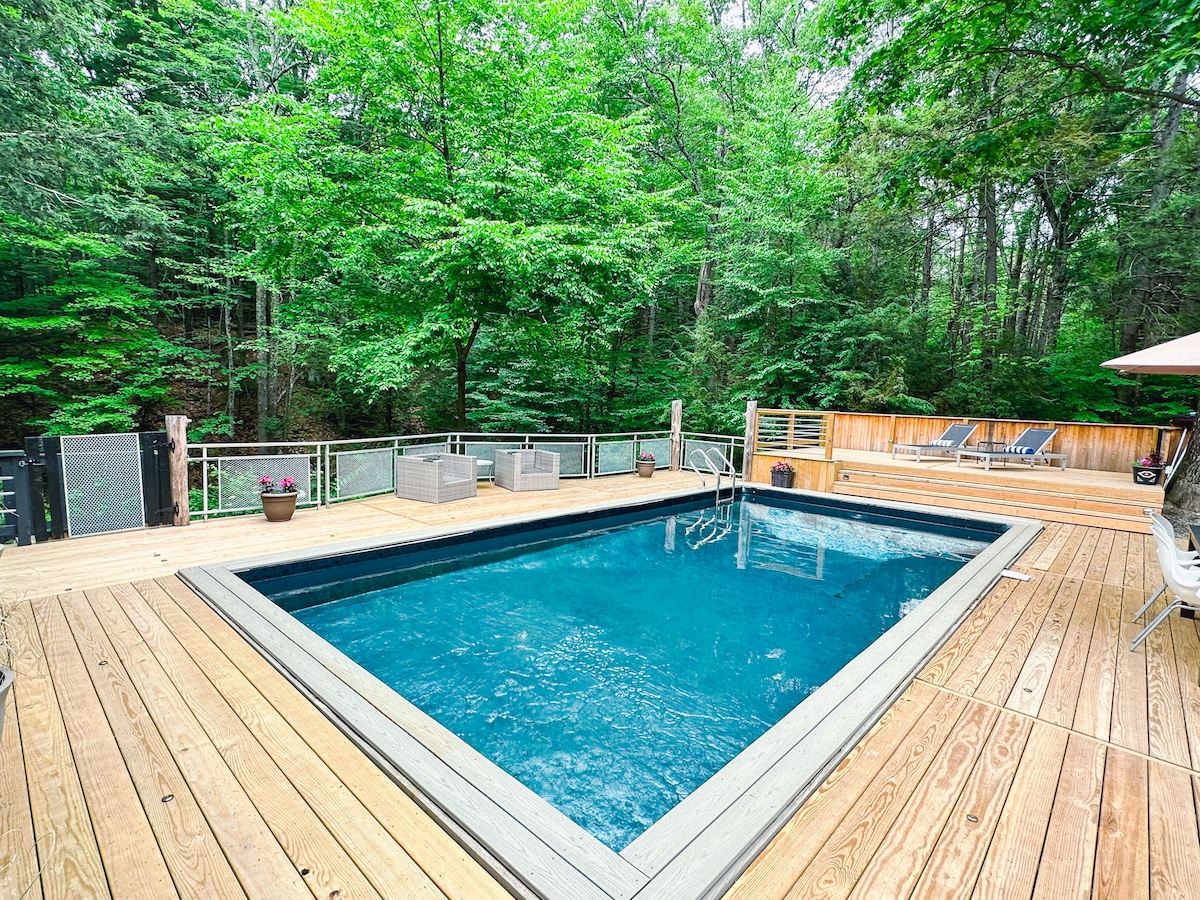 A heated salt-water pool is surrounded by a spacious wooden deck. Comfortable lounge chairs are positioned nearby, and greenery from the surrounding trees creates a private, tranquil environment. The pool's clear blue water contrasts with the natural tones of the deck and landscaping.