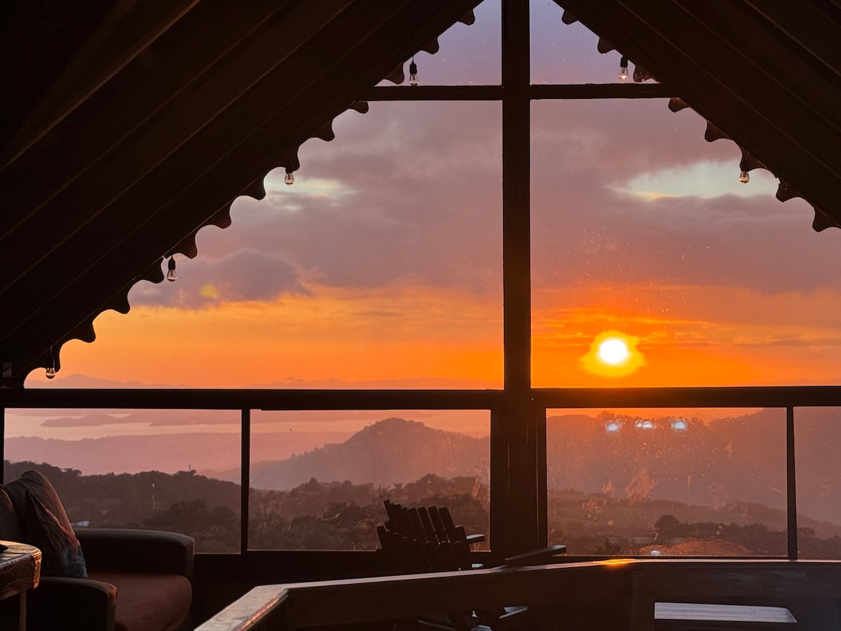 A breathtaking sunset is viewed through large windows, filling the room with warm hues of orange and gold. The silhouette of the mountains and distant hills adds depth to the scene, while hanging light fixtures provide subtle illumination along the wooden ceiling.