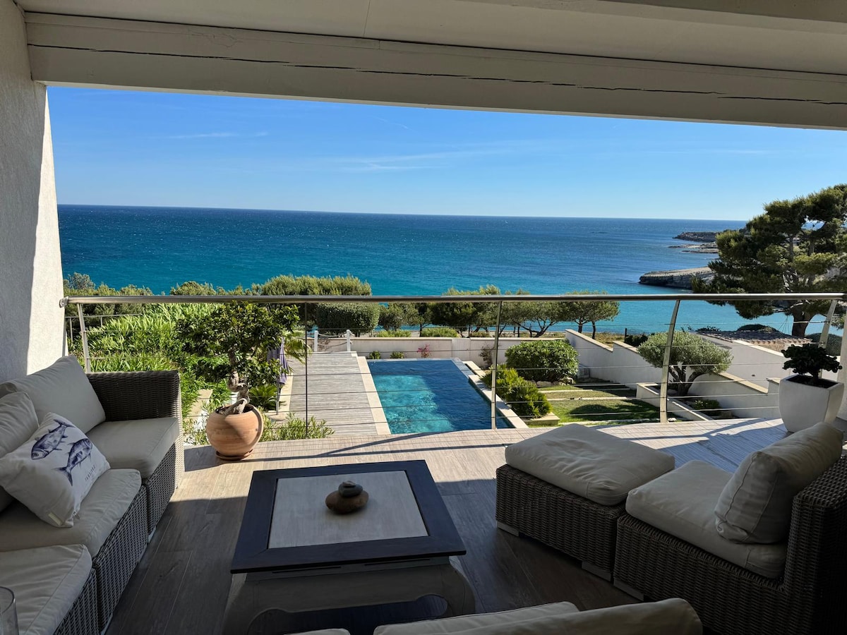 A spacious terrace offers a panoramic view of the ocean, illuminated by natural light. Comfortable seating surrounds a coffee table, inviting relaxation. The scene transitions to a private pool and well-kept gardens, seamlessly blending indoor and outdoor living spaces.