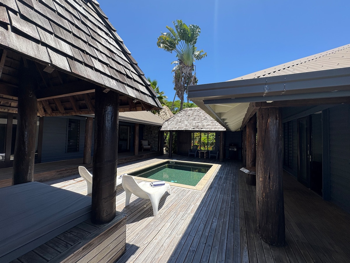 A private courtyard is presented, featuring a marble-tiled plunge pool surrounded by wooden decks. Separate living and sleeping pods are visible, with a thatched roof design. Lush tropical foliage is seen in the background against a clear blue sky, adding to the serene atmosphere of the space.