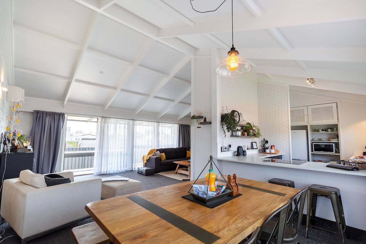 An open plan living and dining area features a spacious wooden table surrounded by metal chairs. A neutral-toned couch is positioned nearby, with a cozy seating arrangement visible in the background. Windows allow natural light to fill the space, creating a bright and airy ambiance.
