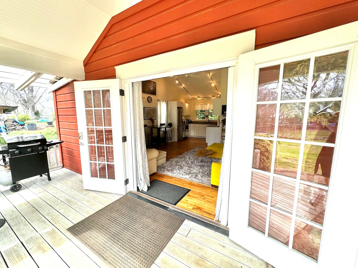 The entrance is framed by double glass doors that open onto a spacious living area. Natural light floods through, highlighting a soft rug and modern furnishings. A grill is positioned on the deck, with a view of the lush yard beyond.