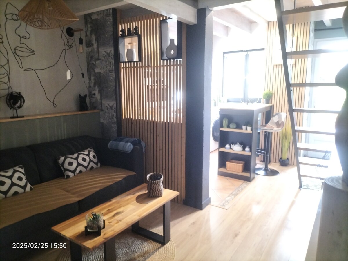 The living area features a comfortable sofa adorned with decorative cushions, complemented by a wooden coffee table. On the wall, decorative accents are arranged, while the light-filled space transitions towards an open kitchen with modern design elements and wooden slatted panels, enhancing the inviting ambiance.