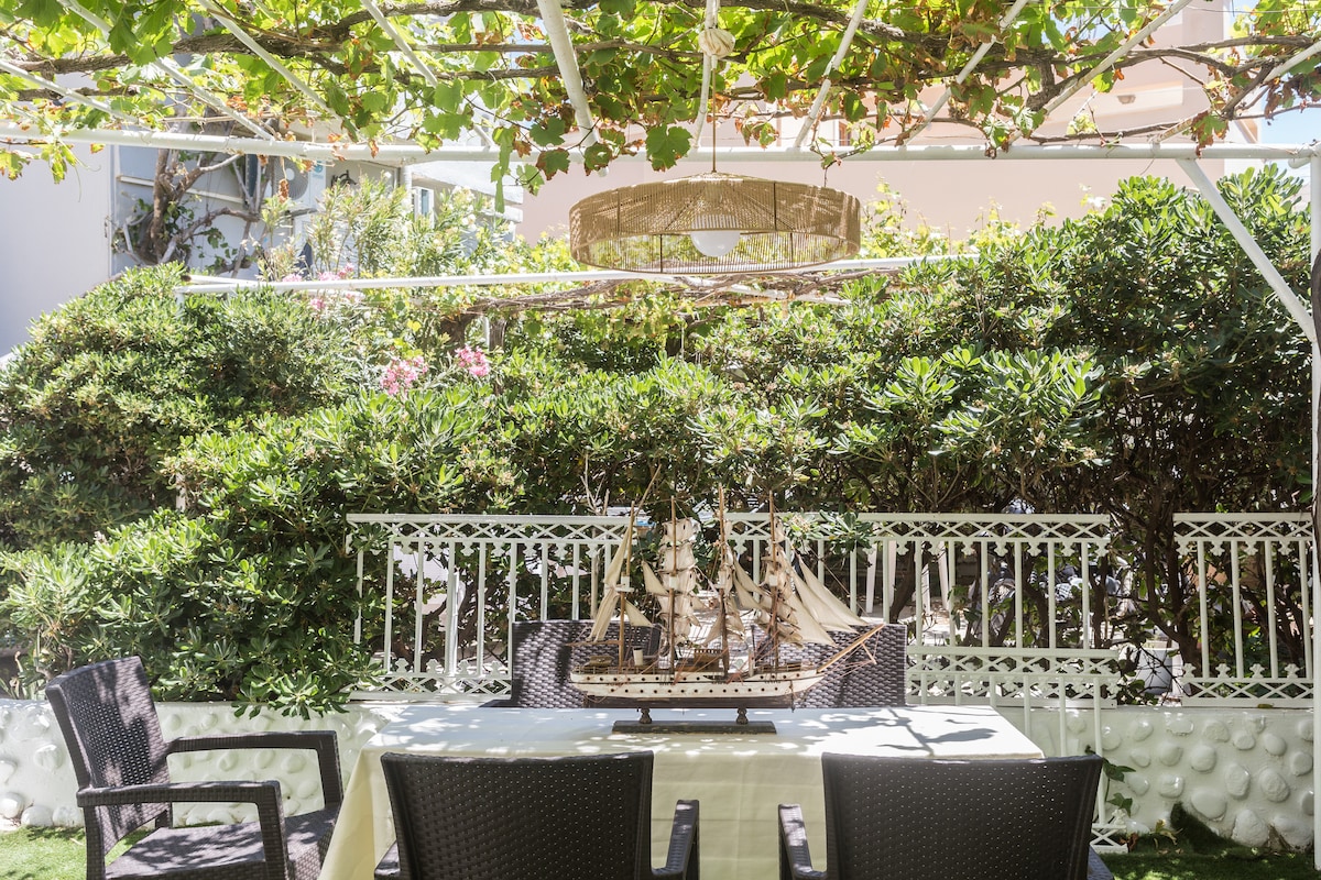 An outdoor dining area is set beneath a shaded pergola, featuring a table surrounded by four wicker chairs. Lush greenery and flowering plants create a natural backdrop, while a decorative ship model is displayed on the table, adding an interesting focal point.