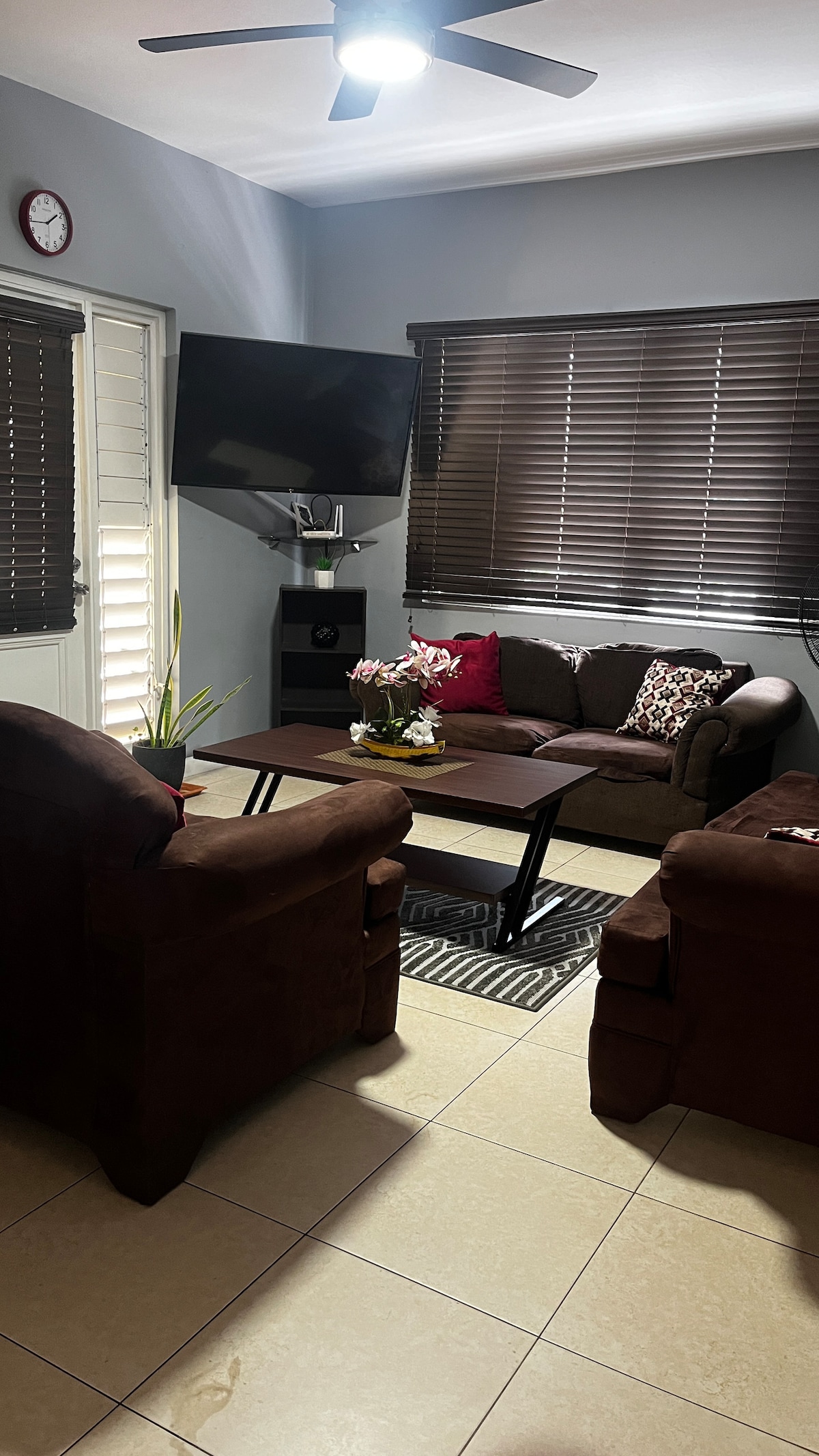 A comfortable living area is presented, featuring a dark brown sofa set positioned around a modern coffee table. A mounted flat-screen television hangs on the wall, while natural light filters through dark window blinds. Decorative plants and a flower arrangement are visible on the table.