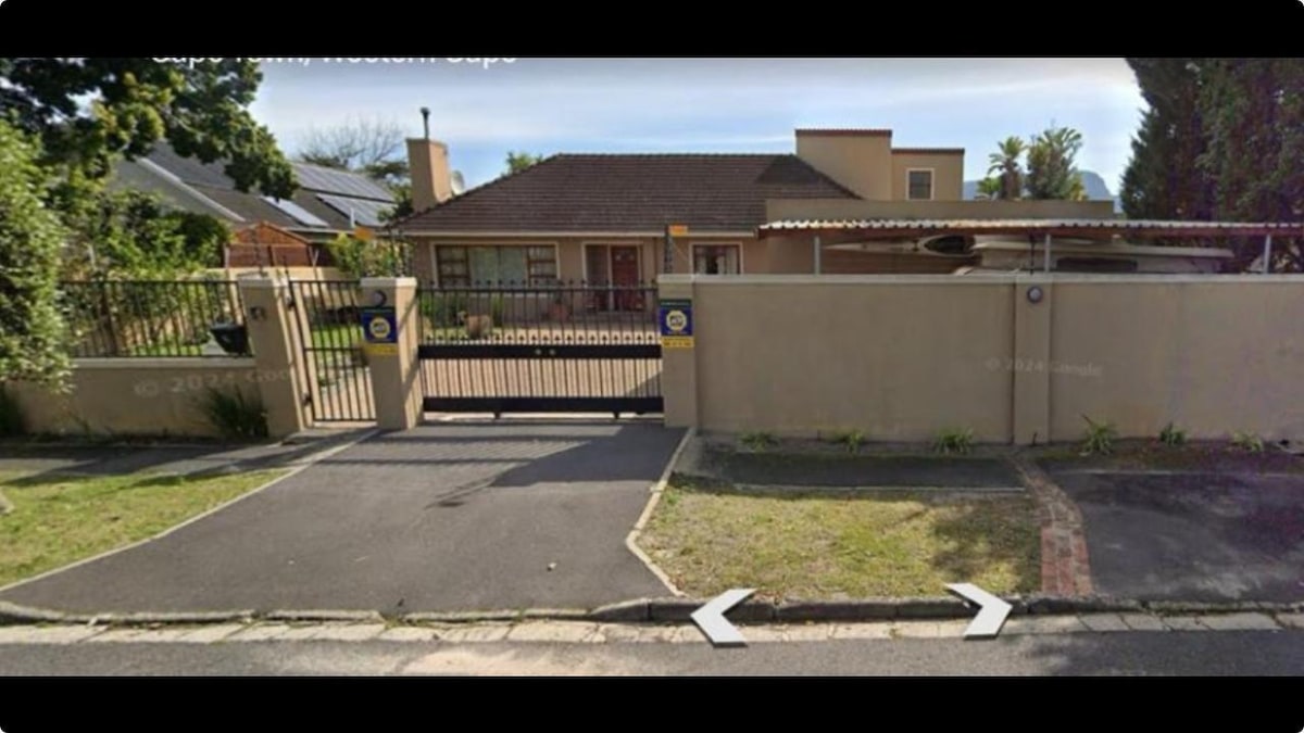 A private entrance to a family home is shown, featuring a fenced yard with a gated driveway. Green shrubs flanking the entrance provide a natural feel, while a shaded porch is visible on the right side of the building.