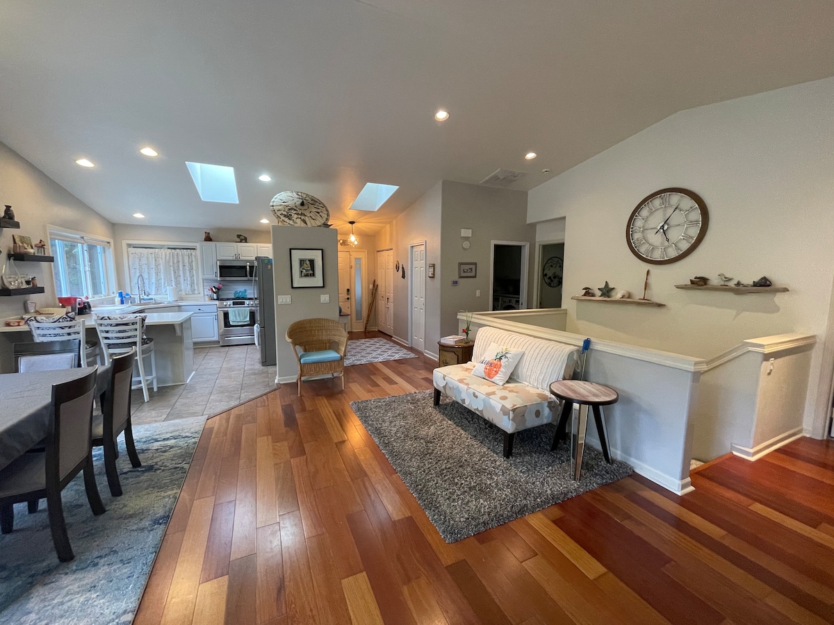 An open-concept living area is illuminated by natural light from skylights. The space features hardwood flooring, a cozy sofa set on a soft area rug, and a dining area with a table. The kitchen is visible in the background, showcasing modern appliances and a casual dining setup.