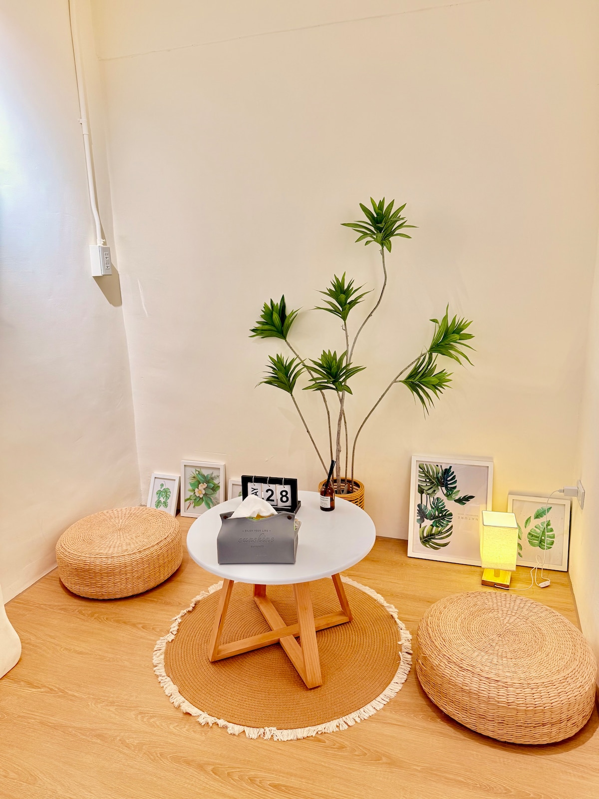 A small seating area features a round white table at the center, surrounded by two woven poufs. A potted plant adds a touch of greenery, while art frames with leaf designs are arranged along the wall. Soft light emits from a standing lamp.