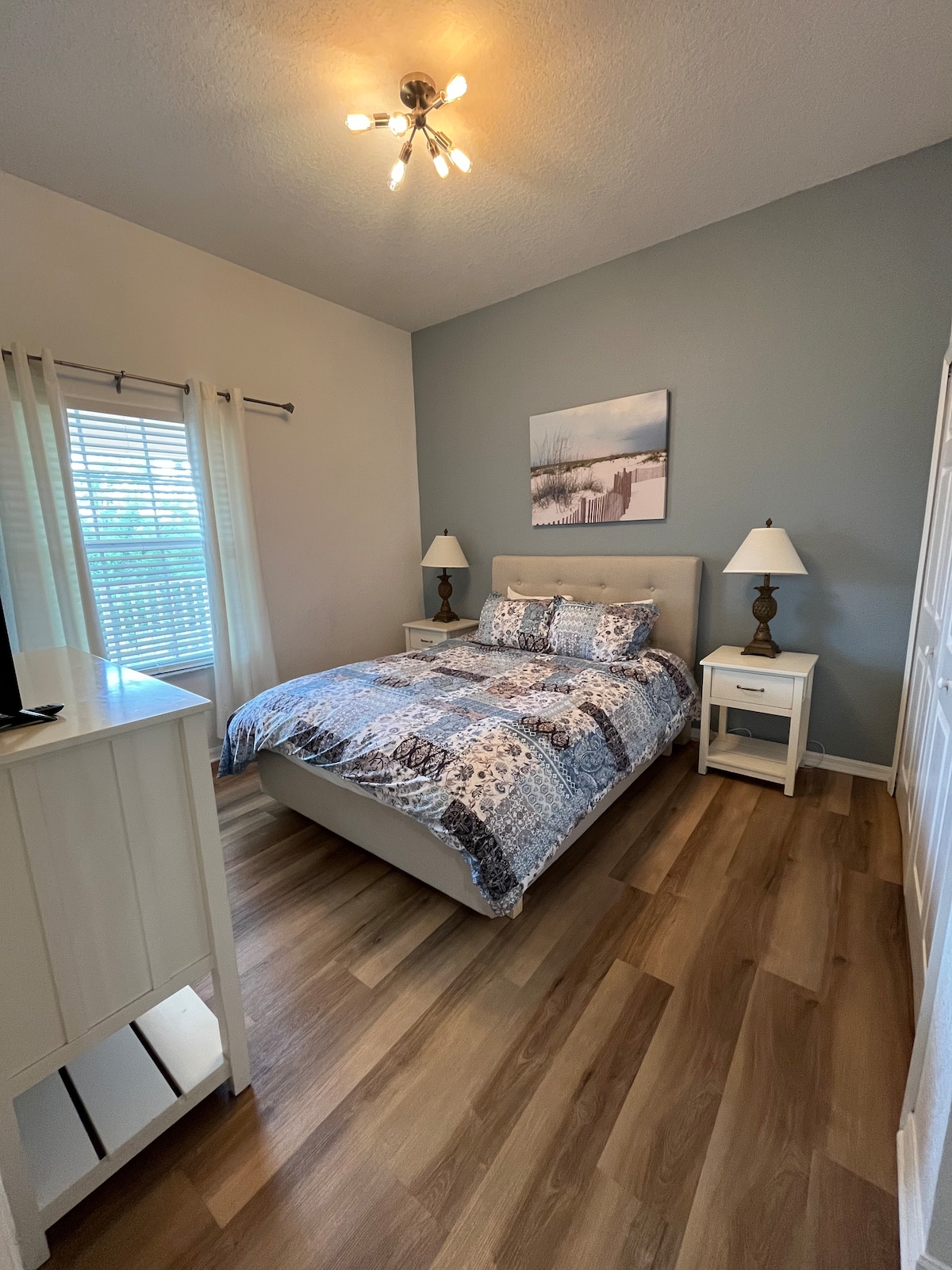 A well-appointed bedroom features a spacious bed dressed in a patterned comforter. Two bedside tables with lamps are positioned on either side of the bed. Natural light enters through a window with blinds, while a piece of artwork hangs above the bed. Light wood flooring enhances the room's appearance.