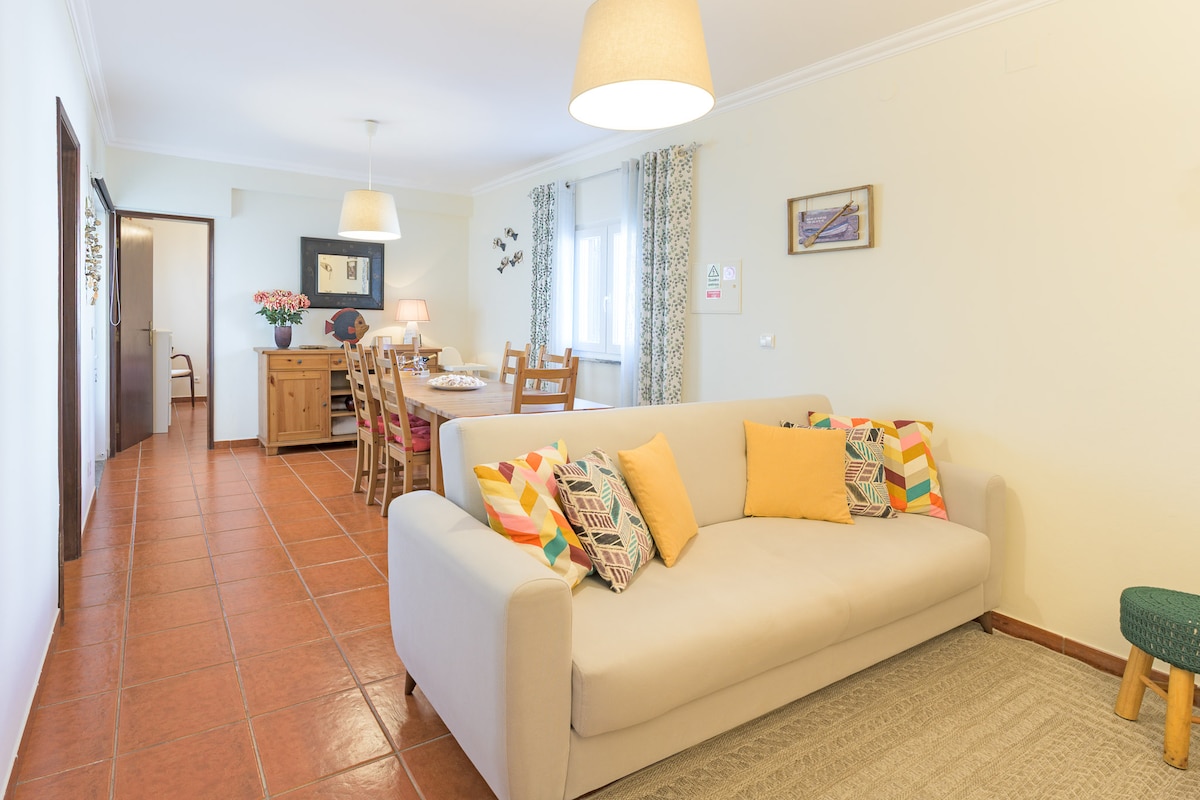 A spacious living area is showcased, featuring a light-colored sofa adorned with decorative cushions. A dining table is visible in the background, surrounded by wooden chairs. Natural light fills the room through large windows, and a soft area rug is placed on the tiled floor.