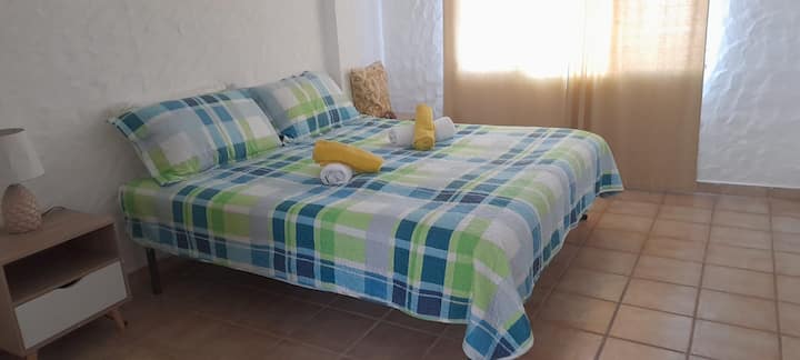 ️ ห้องพักเรียบง่ายและสว่างไสว มีเตียงคู่ ผ้าปูที่นอนลายตารางสีเขียวและฟ้า ม่านที่กรองแสงธรรมชาติ และโต๊ะข้างเตียงพร้อมโคมไฟ ที่พักสะดวกสบายและอบอุ่นสำหรับการพักผ่อน ✨