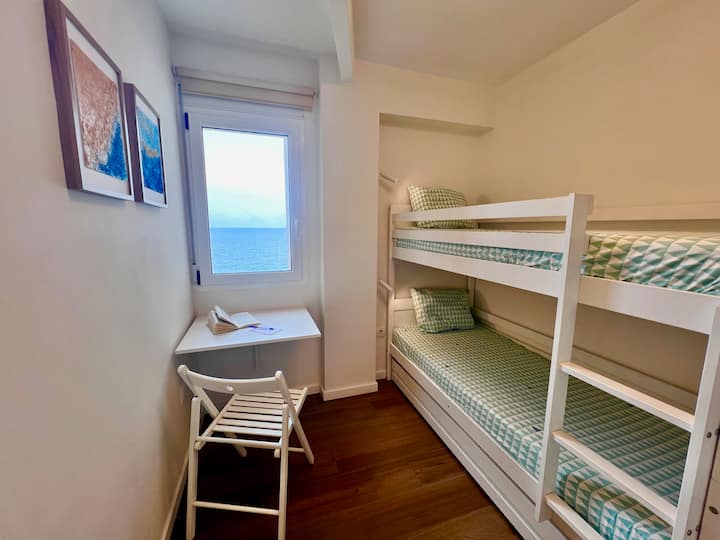 Dormitorio 3 con litera y escritorio abatible bajo la ventana, desde donde se ve el mar. Su tamaño y disposición evocan el camarote de un barco: un rincón acogedor para leer, escribir o dejar volar la imaginación.