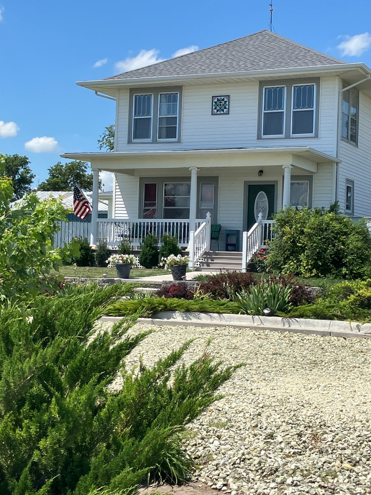 A two-story farmhouse is shown, featuring a welcoming front porch with white railings. Lush greenery and various plants line the path to the entrance, and an American flag gently flutters. Blue skies and fluffy clouds create a serene backdrop.
