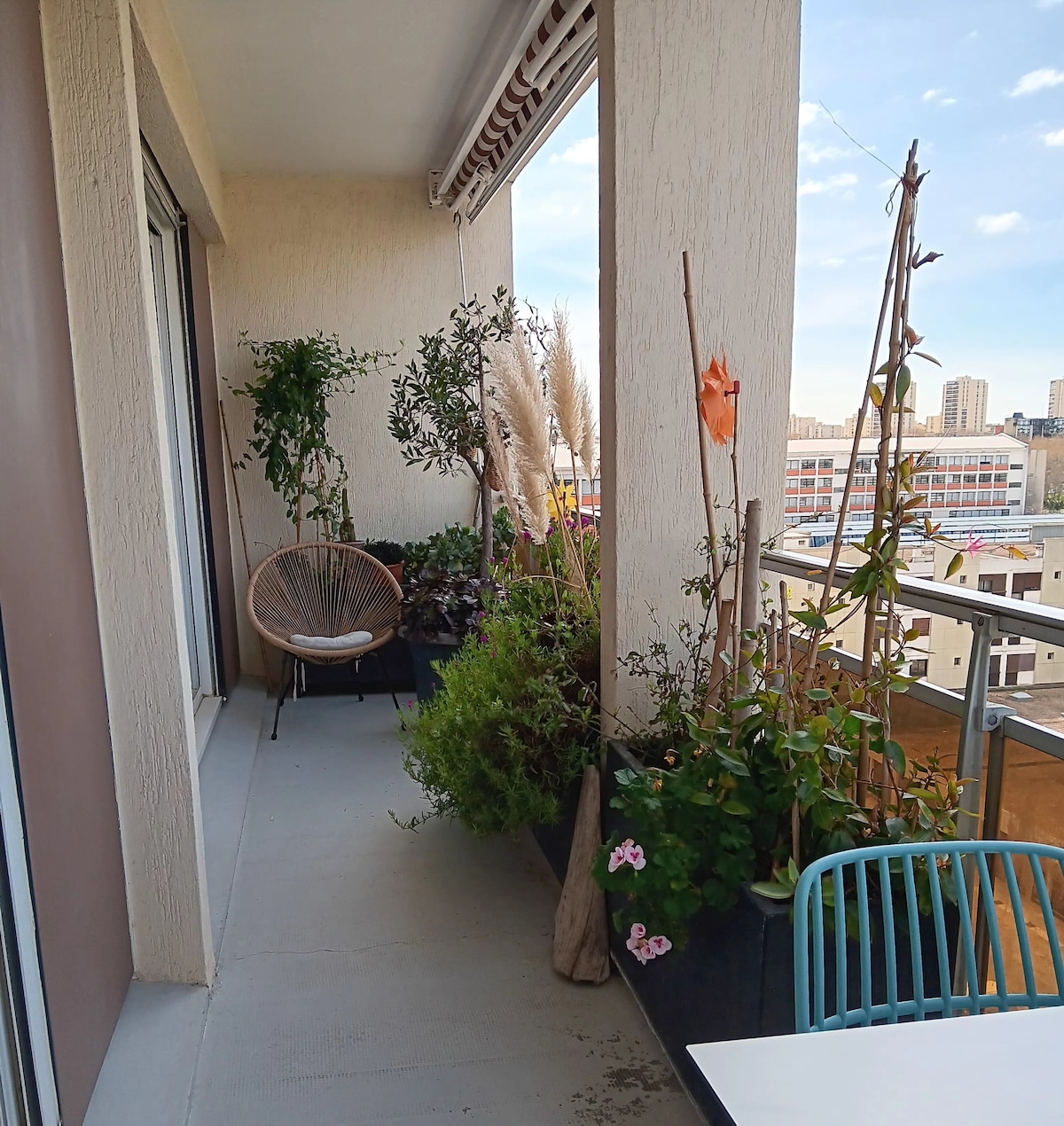 A balcony features a variety of potted plants, including tall grasses and flowering varieties, creating a green atmosphere. Comfortable seating is provided with an Acapulco chair. A small table sits on the balcony, complemented by a backdrop of city buildings and a clear blue sky.
